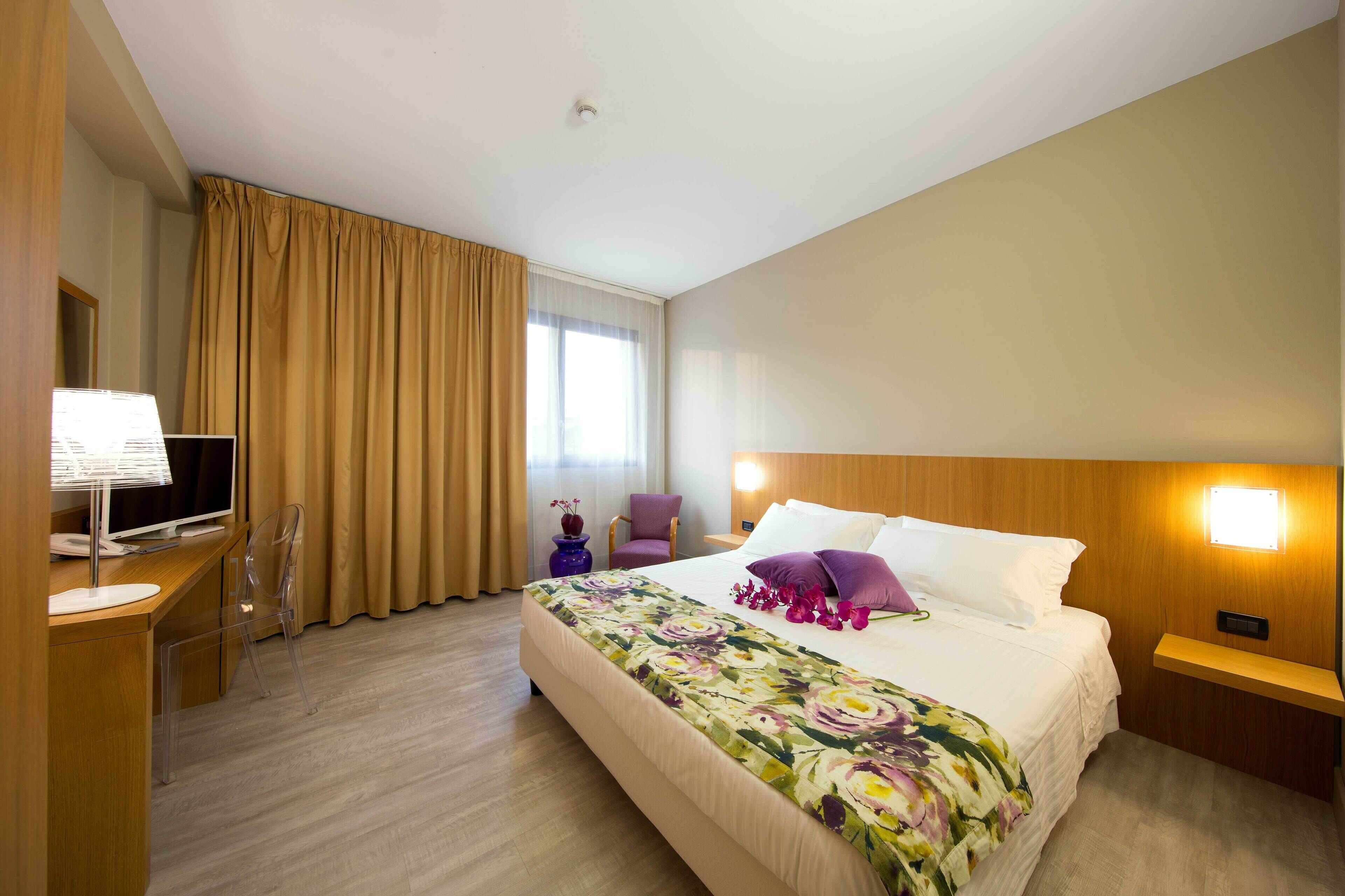A hotel room with a bed, a desk with a computer, a chair, and a nightstand with a lamp.