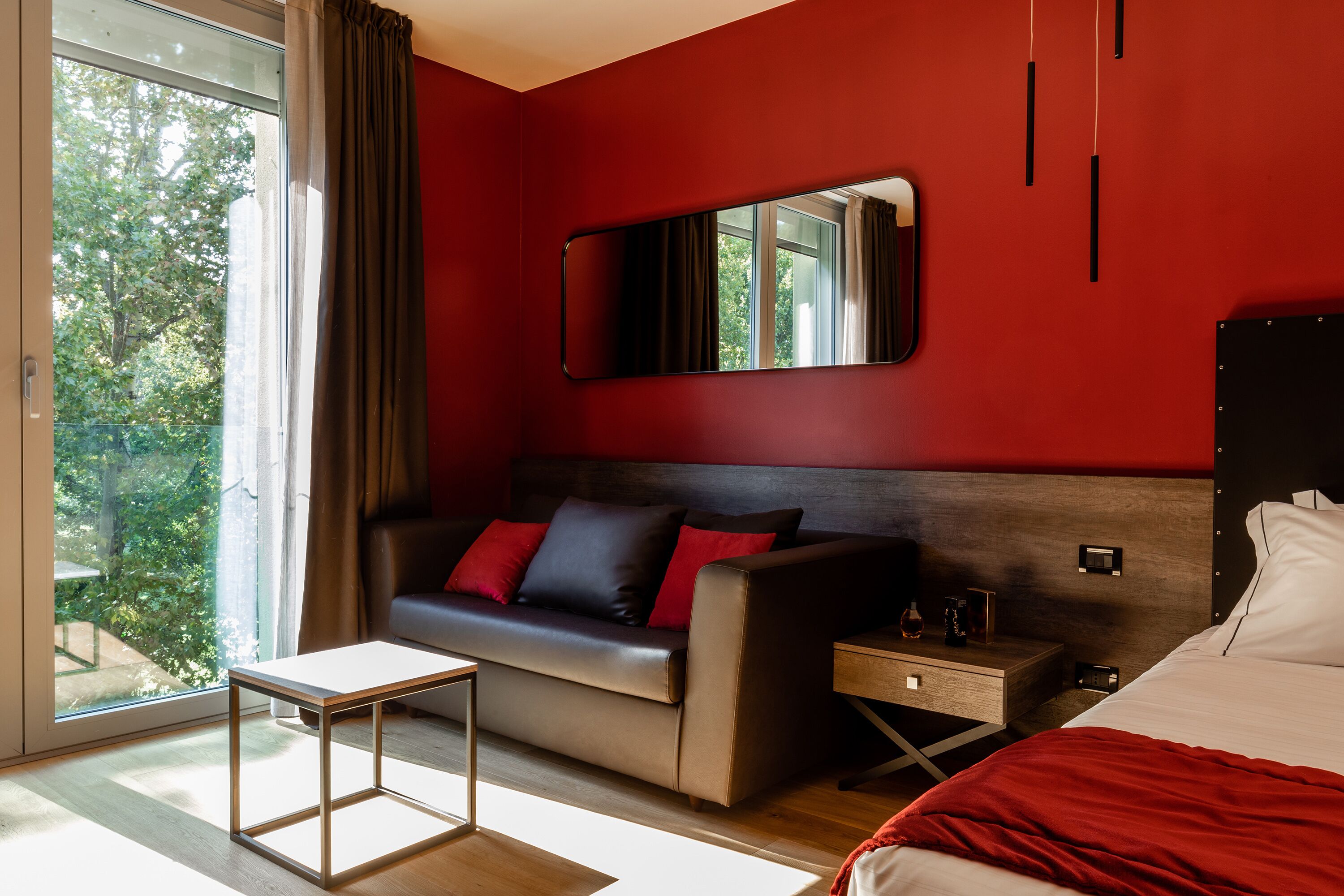 Una habitación de hotel moderna con una pared de acento roja, un sofá con cojines rojos, una mesita de noche, una mesilla auxiliar y una cama con una manta roja, además de un ventanal con vistas a árboles.