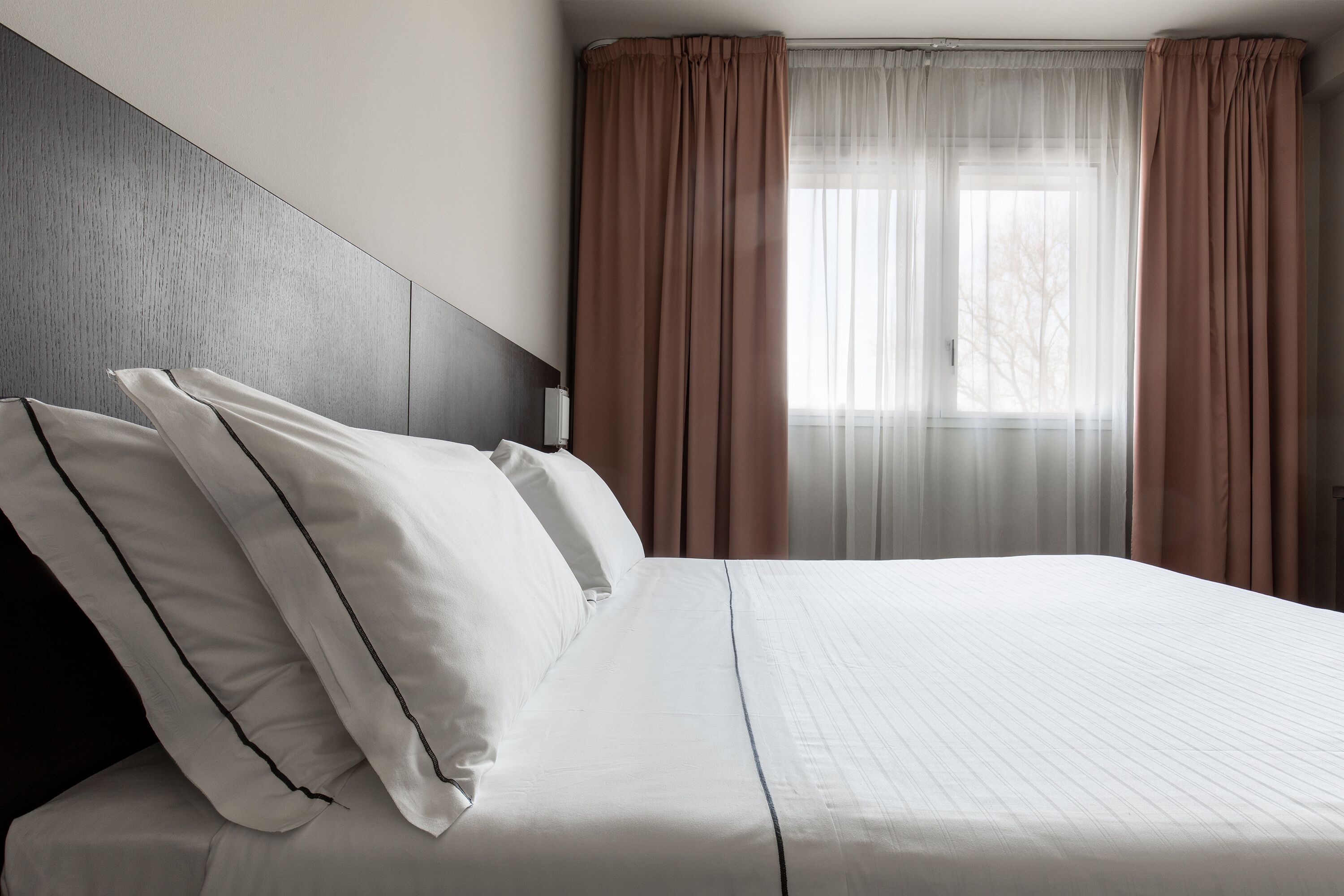A neatly made bed with white linens and black piping, a wooden headboard, and a window with sheer white curtains and brown drapes.