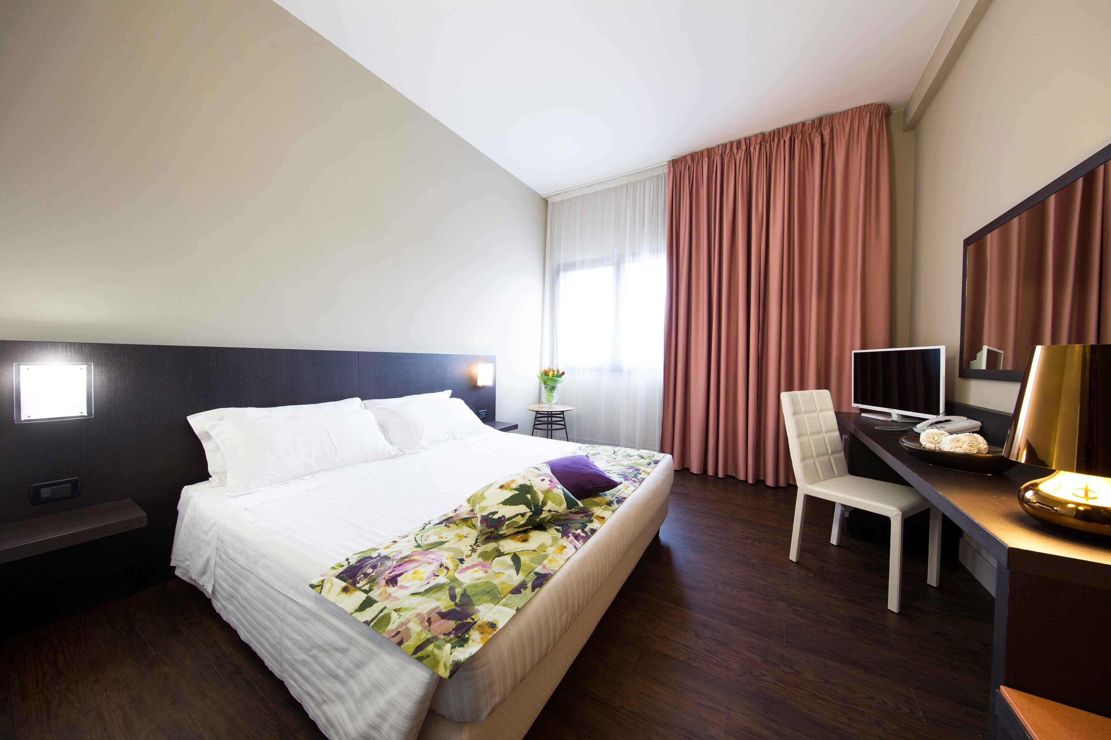 A hotel room with a bed, a desk with a computer, a chair, a TV, and a lamp.
