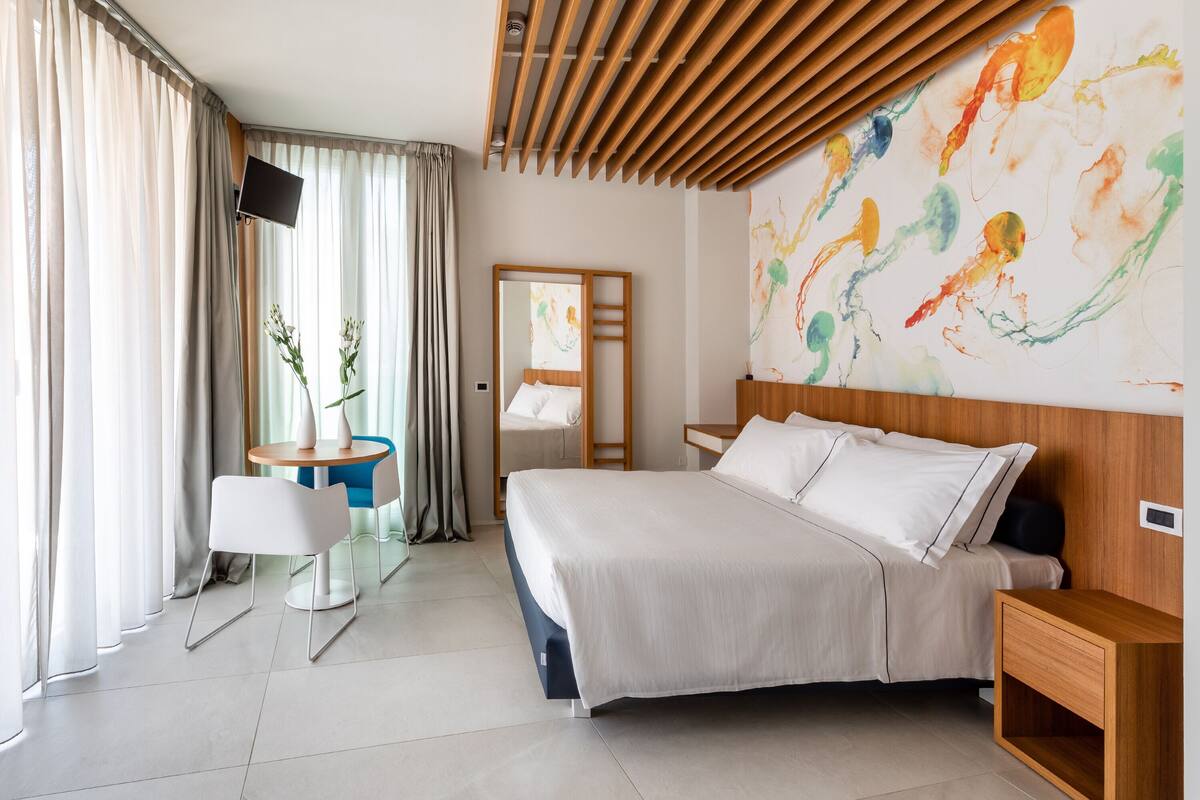 Ein modernes Hotelzimmer mit einem großen Bett, einem Schreibtisch mit Stuhl, einem an der Wand montierten Fernseher und einem großen abstrakten Gemälde.