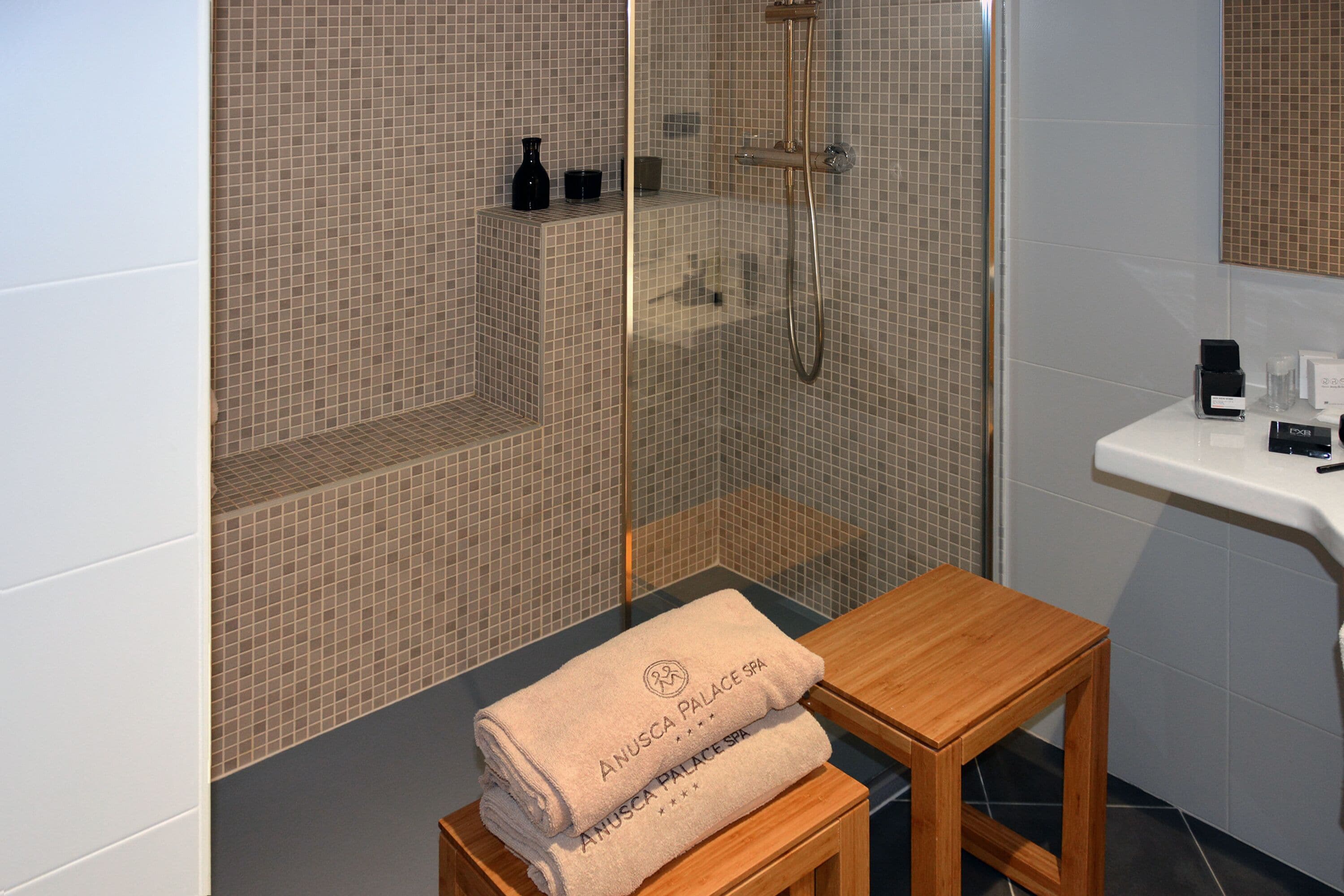 A bathroom with a glass-enclosed shower, a wooden stool, and a sink with toiletries.