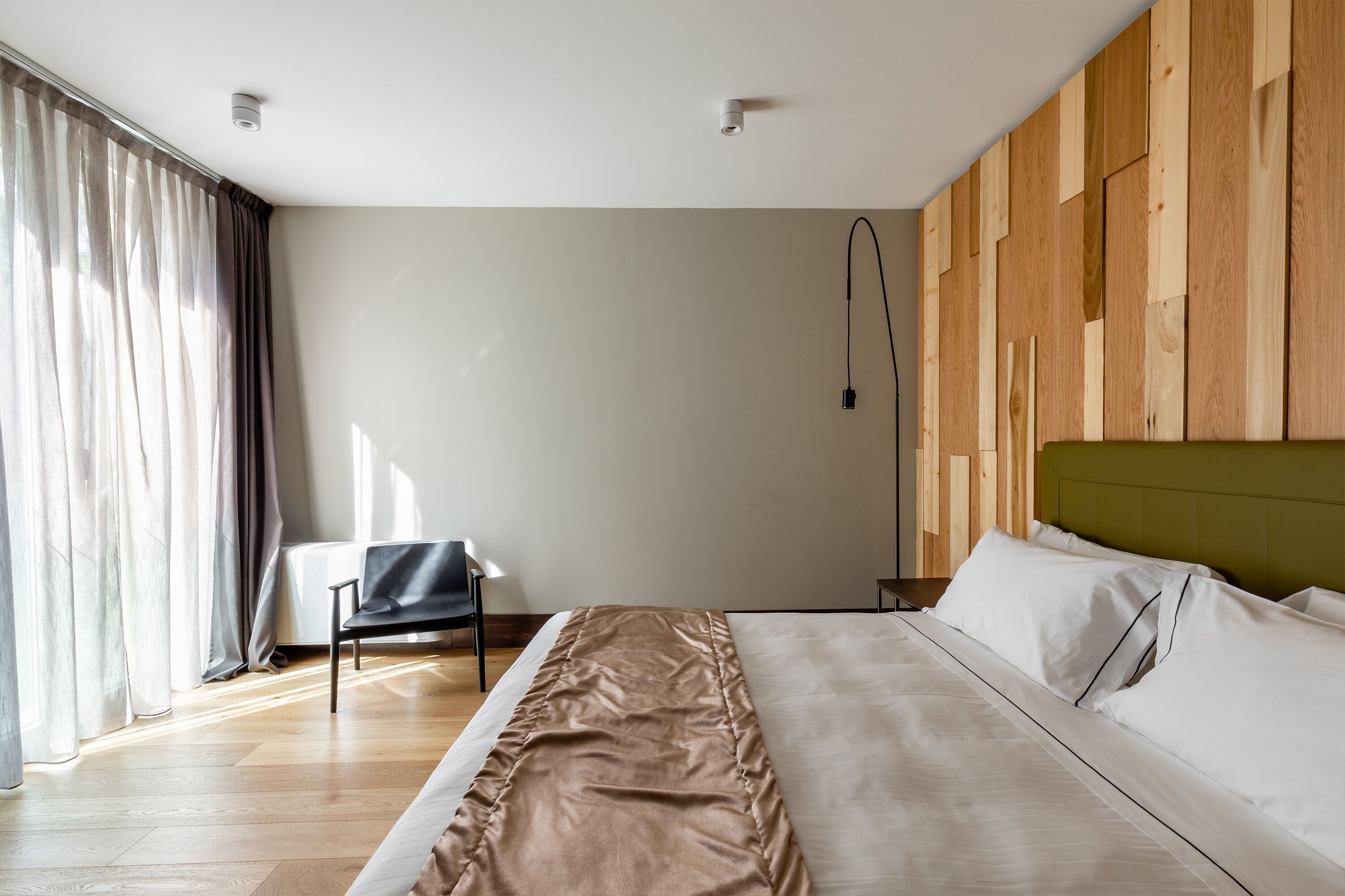 A modern hotel room with a large bed, wooden panel wall, and a chair by the window.