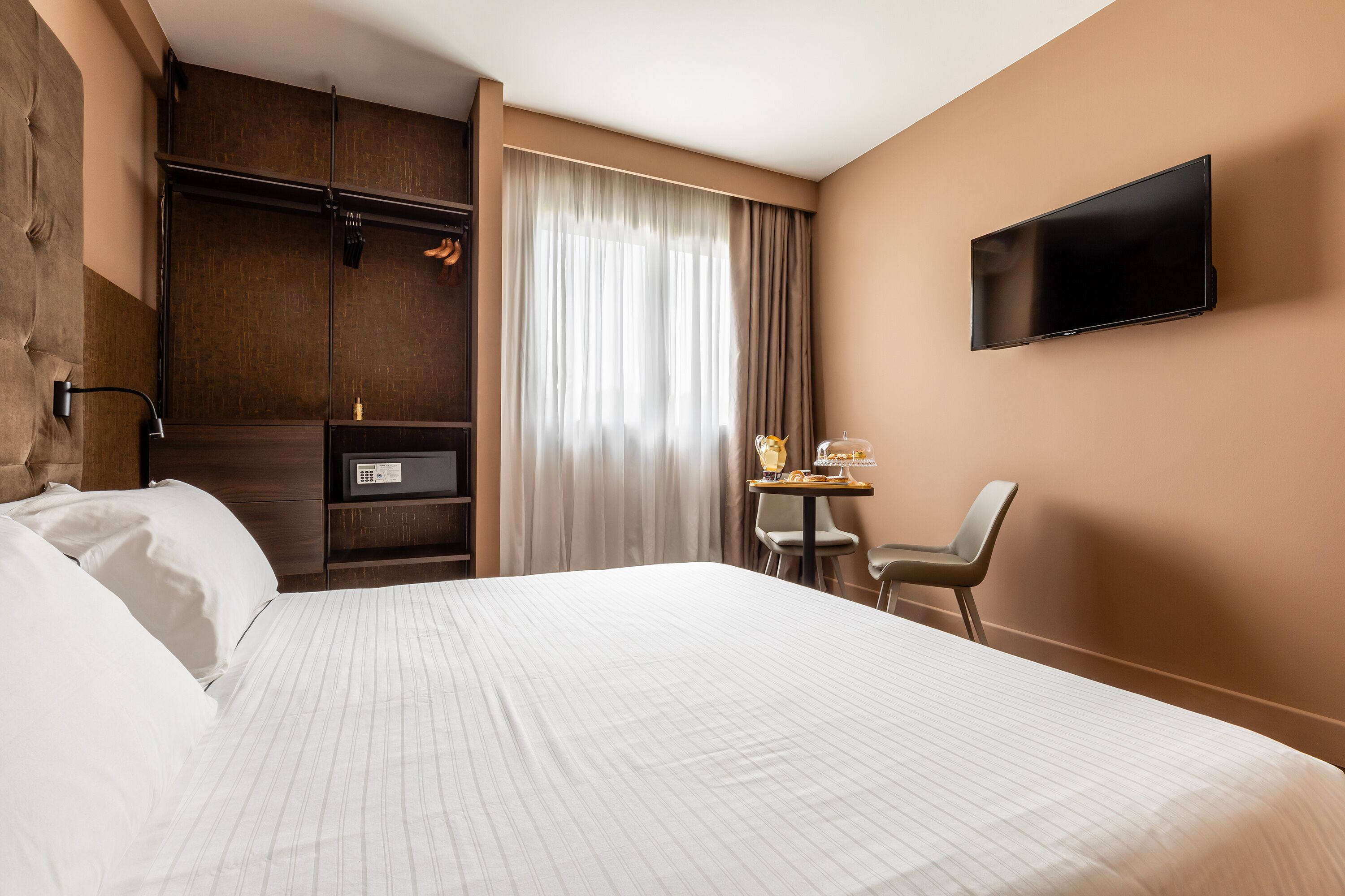 Habitación de hotel con una cama grande, un escritorio con silla, televisor montado en la pared y una ventana con cortinas.