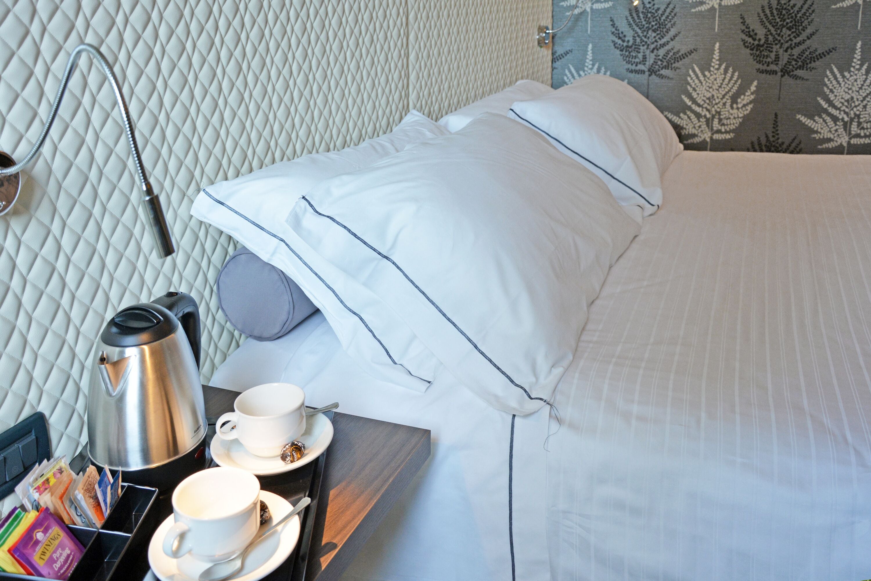 Una cama bien hecha con sábanas y almohadas blancas, una mesita de noche con cafetera, tazas y una bandeja con libros y una tetera.