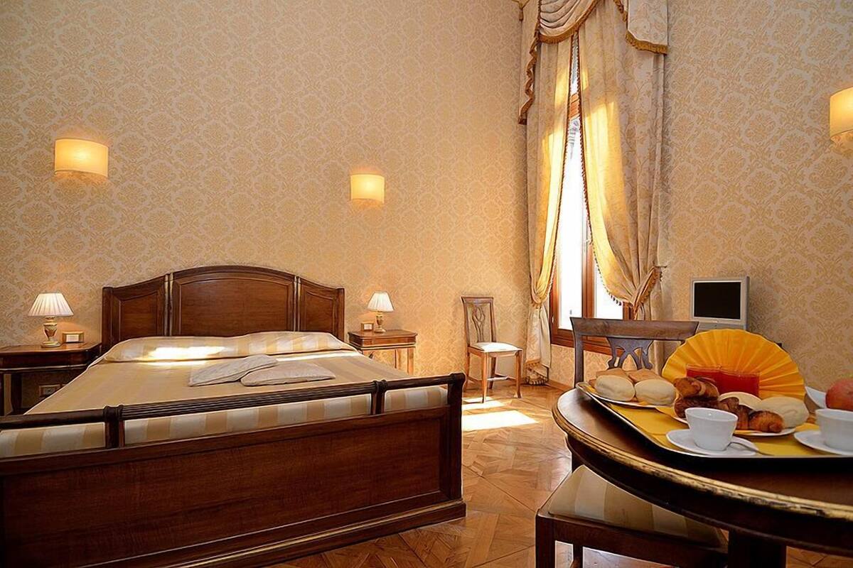 A hotel room with a large bed, two bedside lamps, a small table with breakfast items, and a chair.