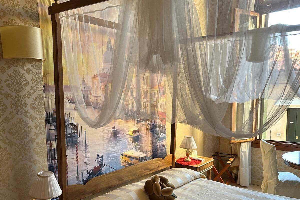 A hotel room with a large bed, a nightstand with a lamp, a chair, and a view of a cityscape through sheer curtains.
