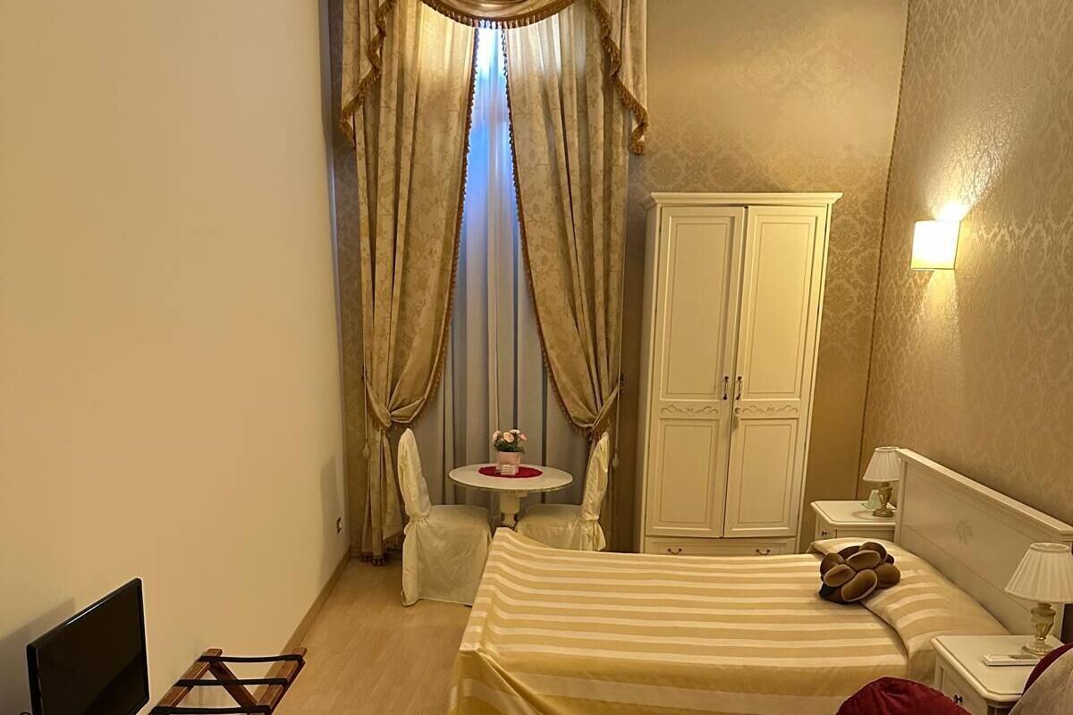 A hotel room with a bed, a desk with a computer, a chair, a small table with flowers, a wardrobe, and a decorative carpet.