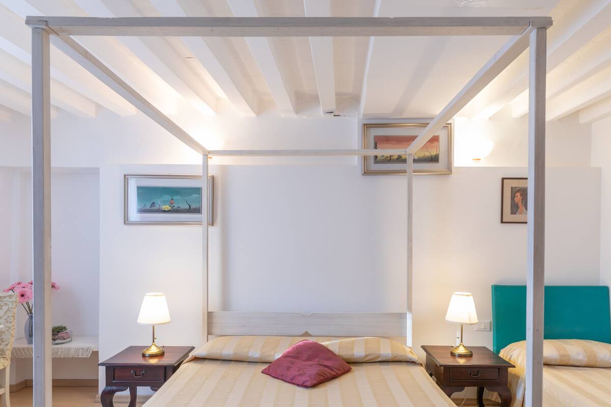 A four-poster bed with a headboard, two bedside tables with lamps, and a window with a view of the sky.