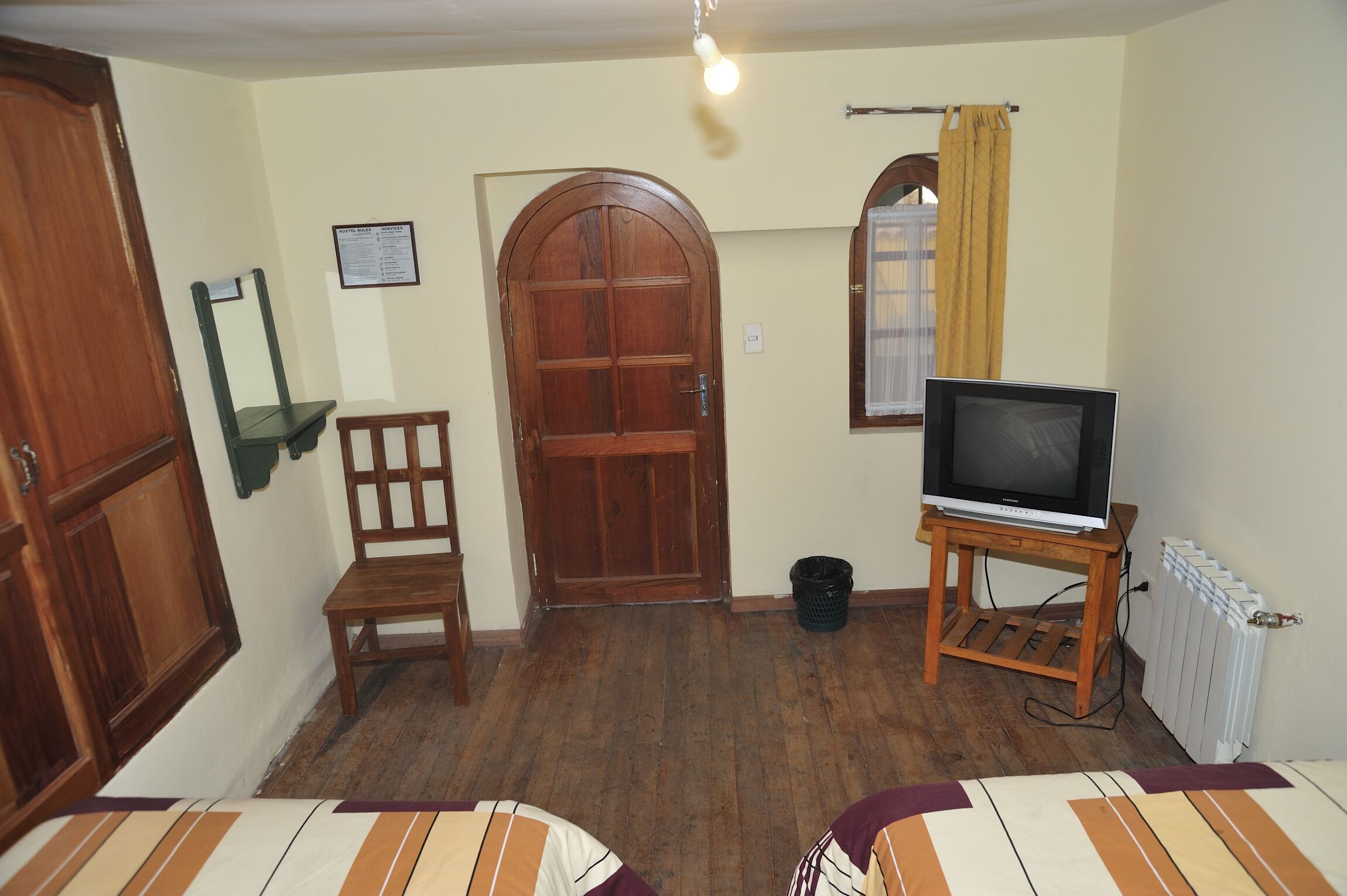 A room with a wooden door, a chair, a television on a stand, a radiator, and a window with a curtain.