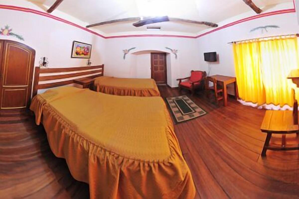 A room with two beds, a wooden floor, a desk, a chair, a television, and a window with curtains.