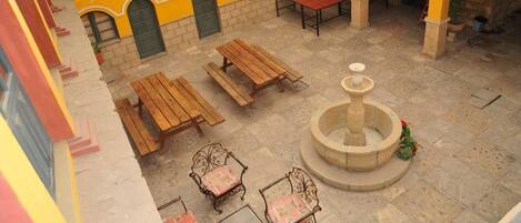 Une cour extérieure avec des bancs en bois, une fontaine et un bâtiment aux murs jaunes et aux fenêtres vertes.