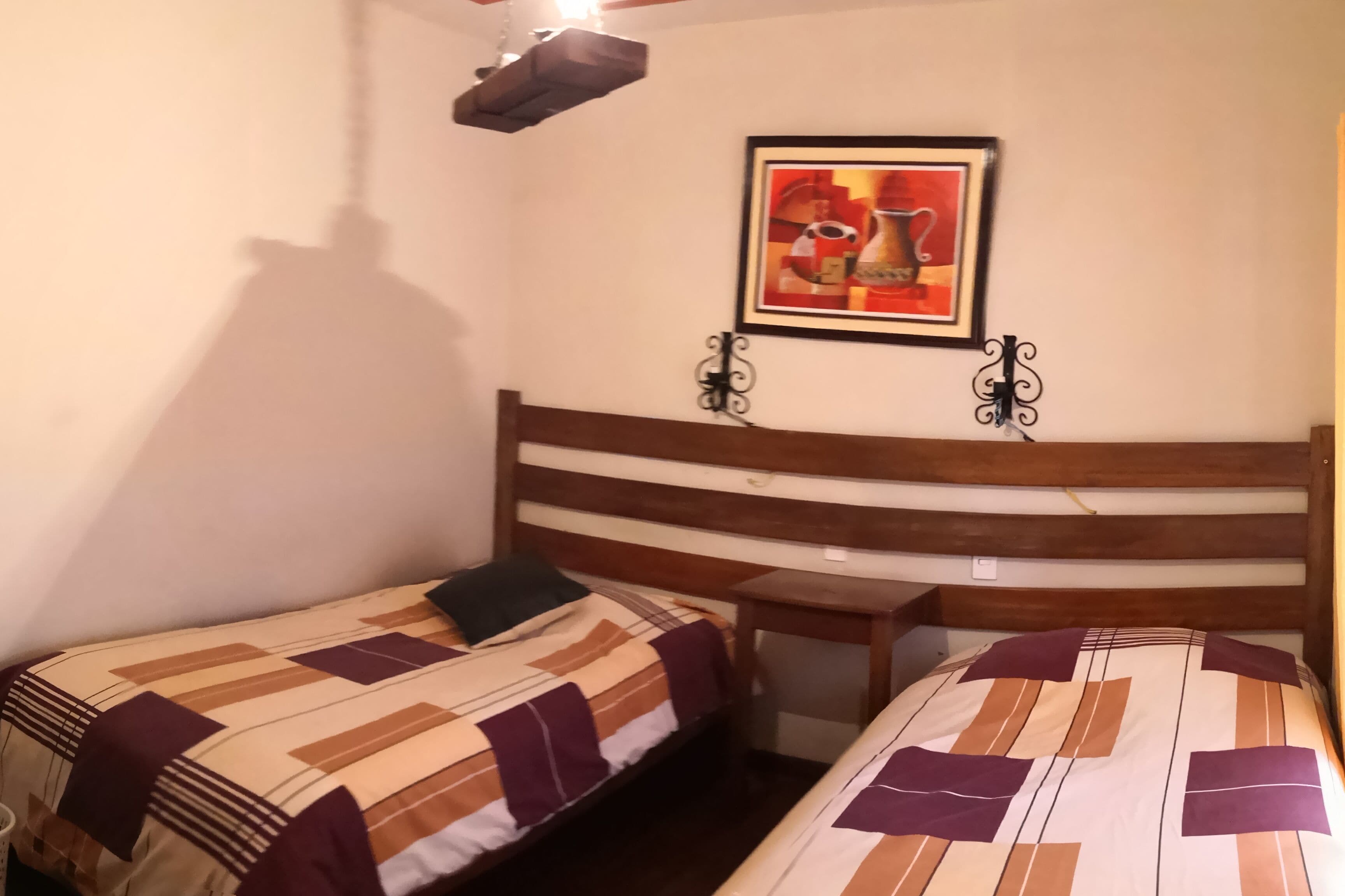 A small, cozy room with two single beds, a wooden headboard, a small table, and a framed picture on the wall.