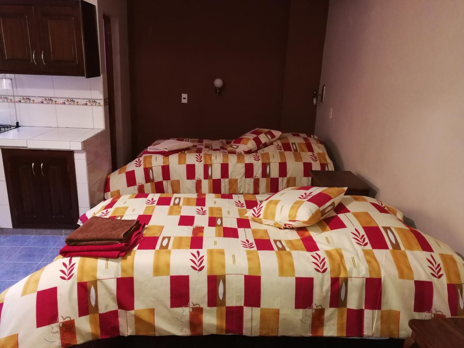 A bedroom with a checkered bedspread, a brown blanket, and a pillow. There is a small bedside table and a wall-mounted light.
