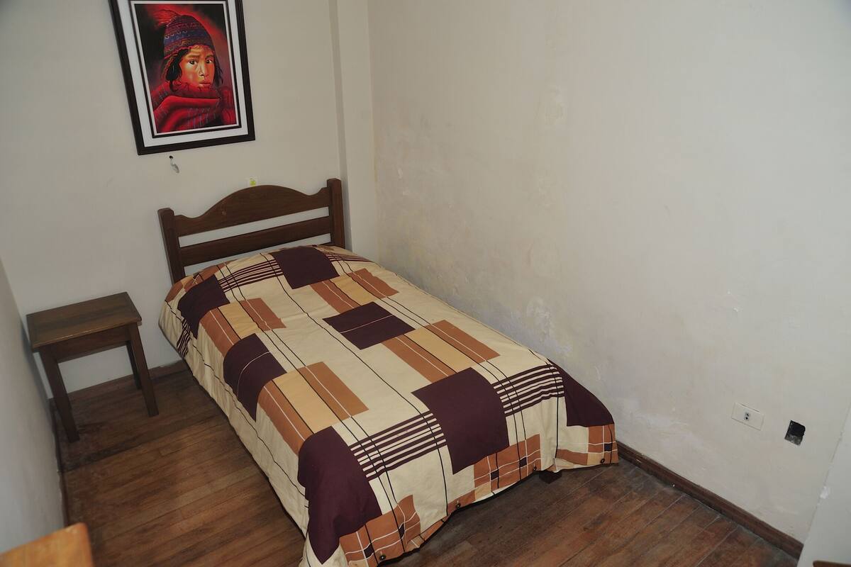 A small, single-bed room with a wooden headboard, a small wooden nightstand, and a plain white wall.