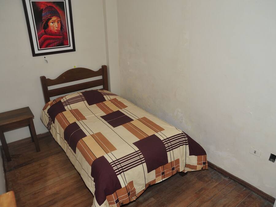 A small, single-bed room with a wooden headboard, a small wooden nightstand, and a plain white wall.