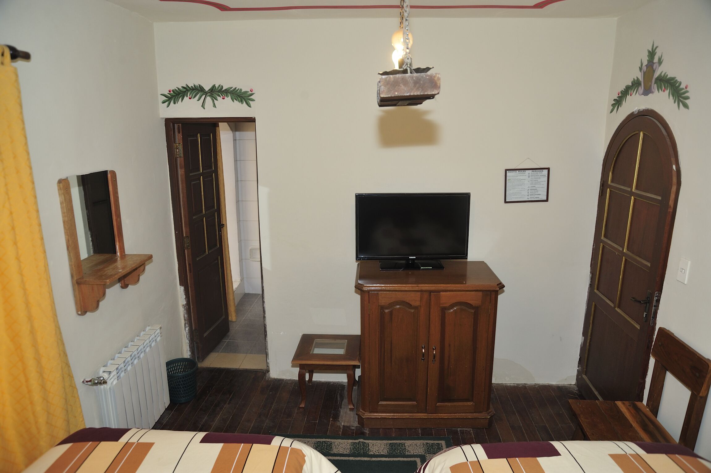 Una habitación con una cama, un televisor sobre un mueble de madera, un radiador, una puerta de madera y una mesita.