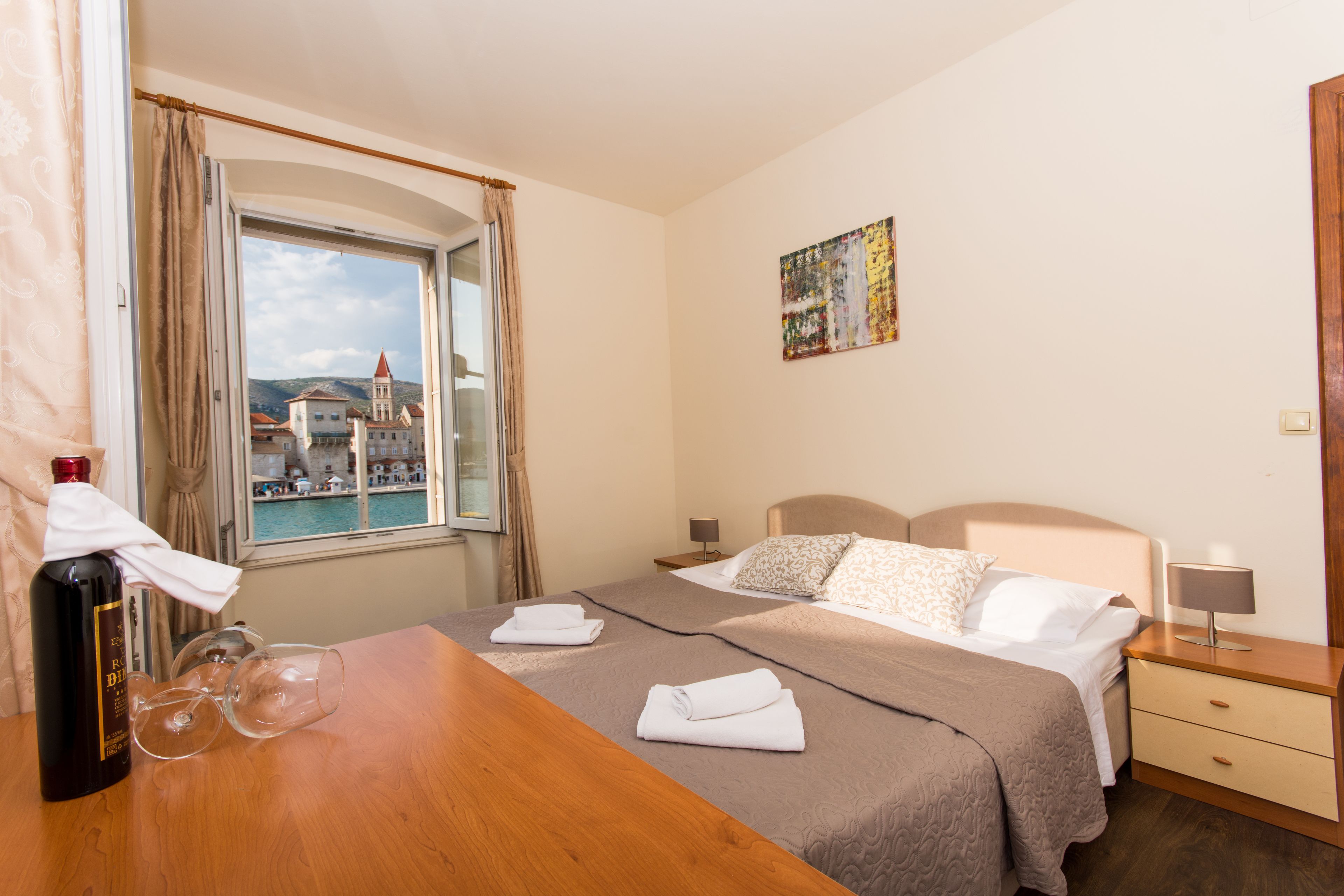 A hotel room with a bed, a wooden table with a bottle and glasses, a nightstand with a lamp, and a window with a view of buildings and water.