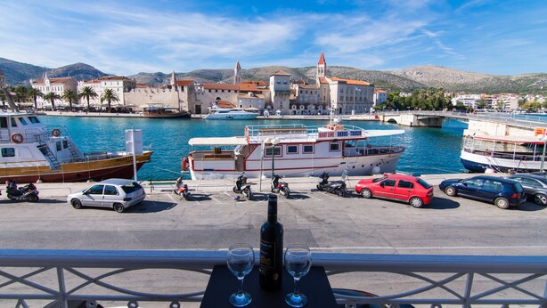 A marina with boats docked, a table with two glasses and a bottle, and a view of buildings and hills in the background. - Vila Liza (Trogir)