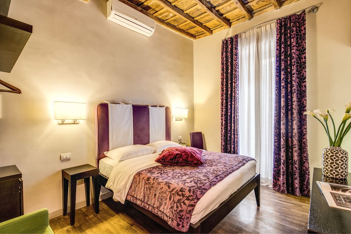 A bedroom with a wooden ceiling, a bed with a purple headboard, a bedside table, a nightstand, a green chair, and a window with purple curtains.