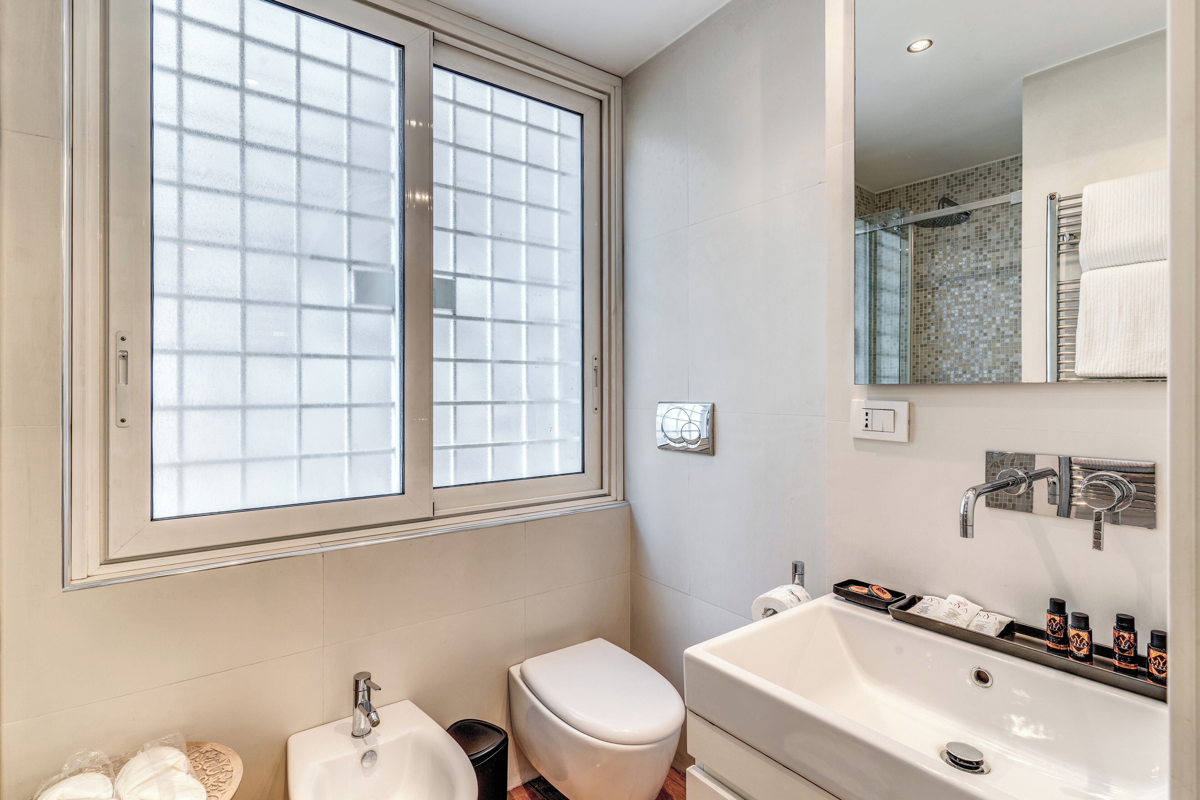 A bathroom with a toilet, sink, and bathtub. There is a window with frosted glass and a mirror above the sink.