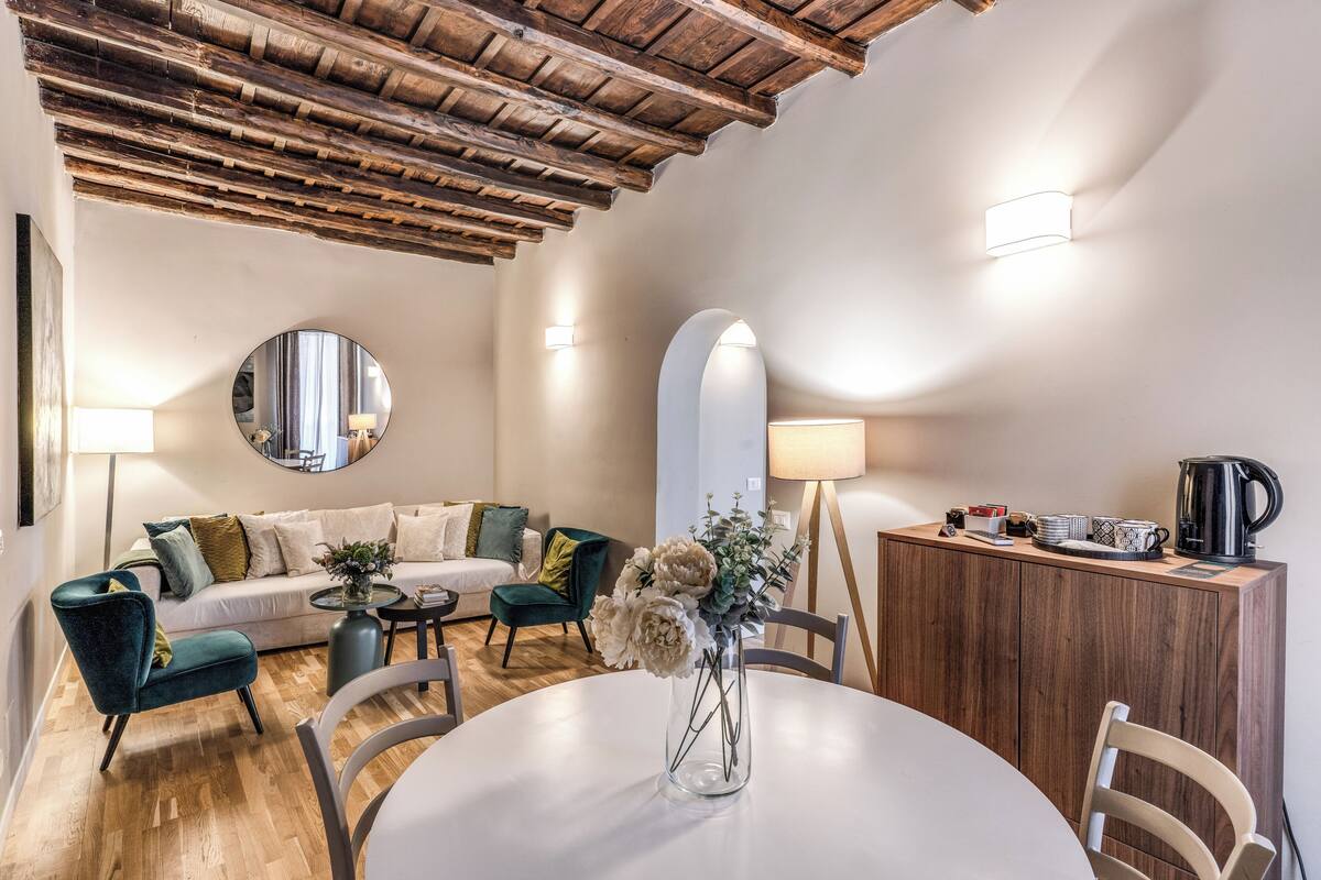 A modern living room with a wooden ceiling, a round mirror, a sofa with cushions, a dining table with chairs, and a cabinet with a kettle.