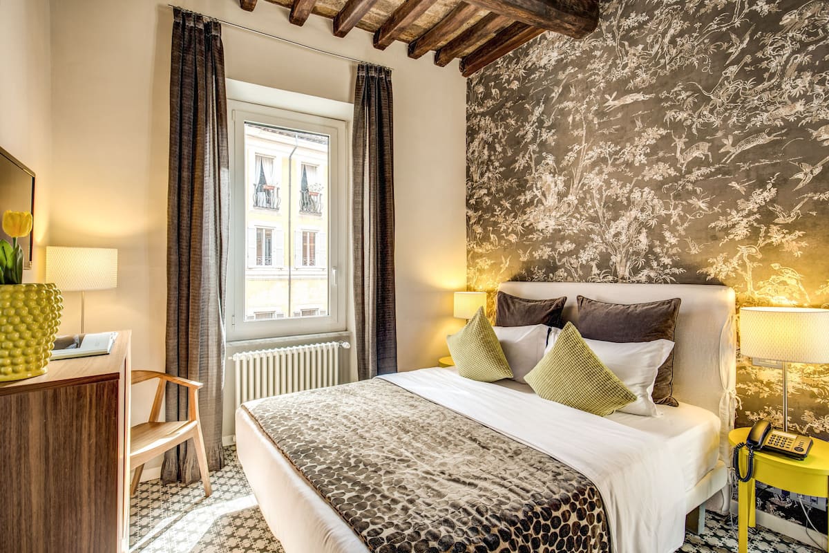 A hotel room with a bed, a desk, a chair, a window with curtains, a radiator, and a patterned wall.