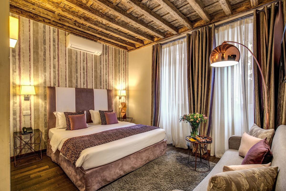 A hotel room with a large bed, a sofa, a small table with a vase, and a mirror.