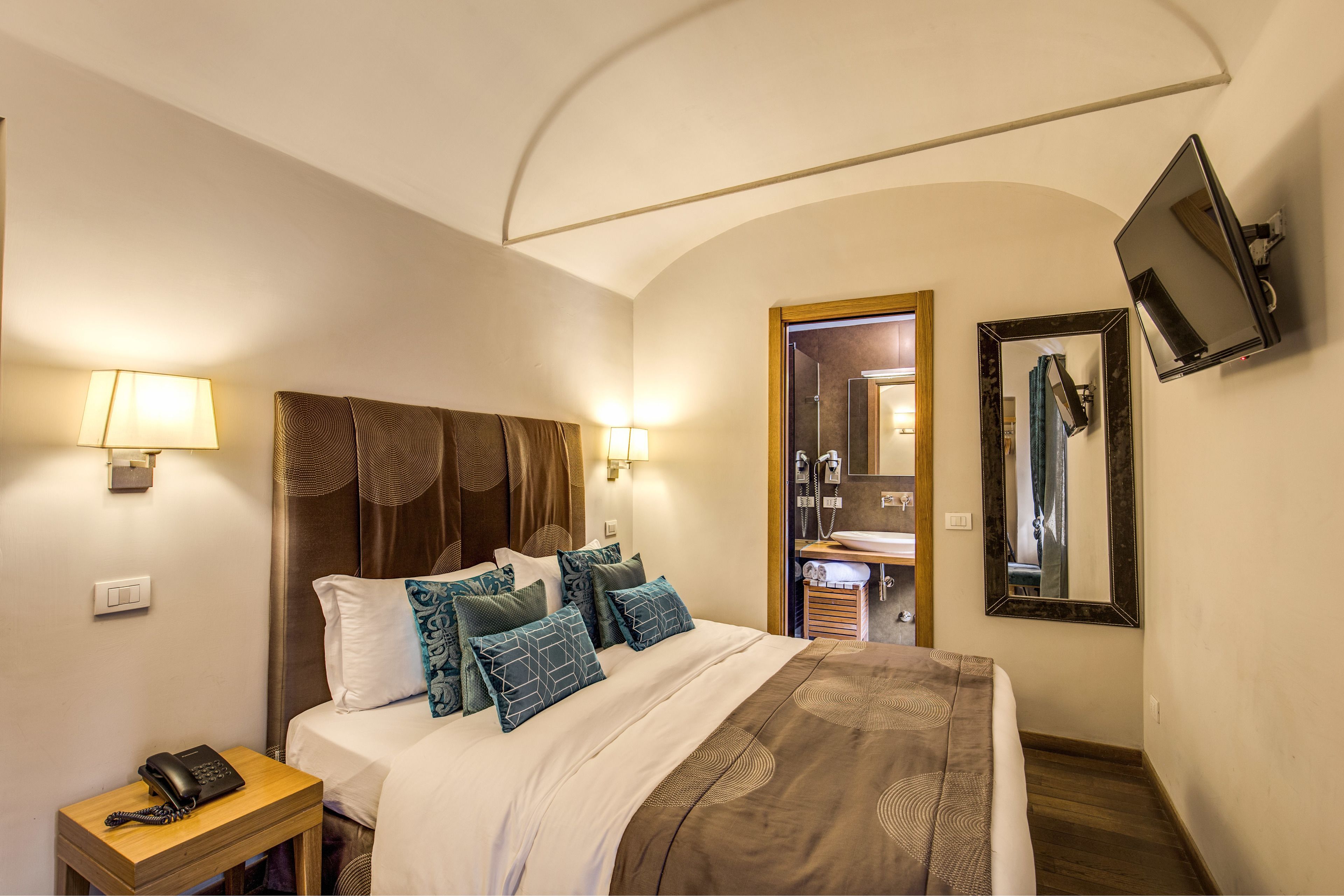 A hotel room with a bed, bedside lamps, a telephone, a mirror, and a bathroom visible through an open door.