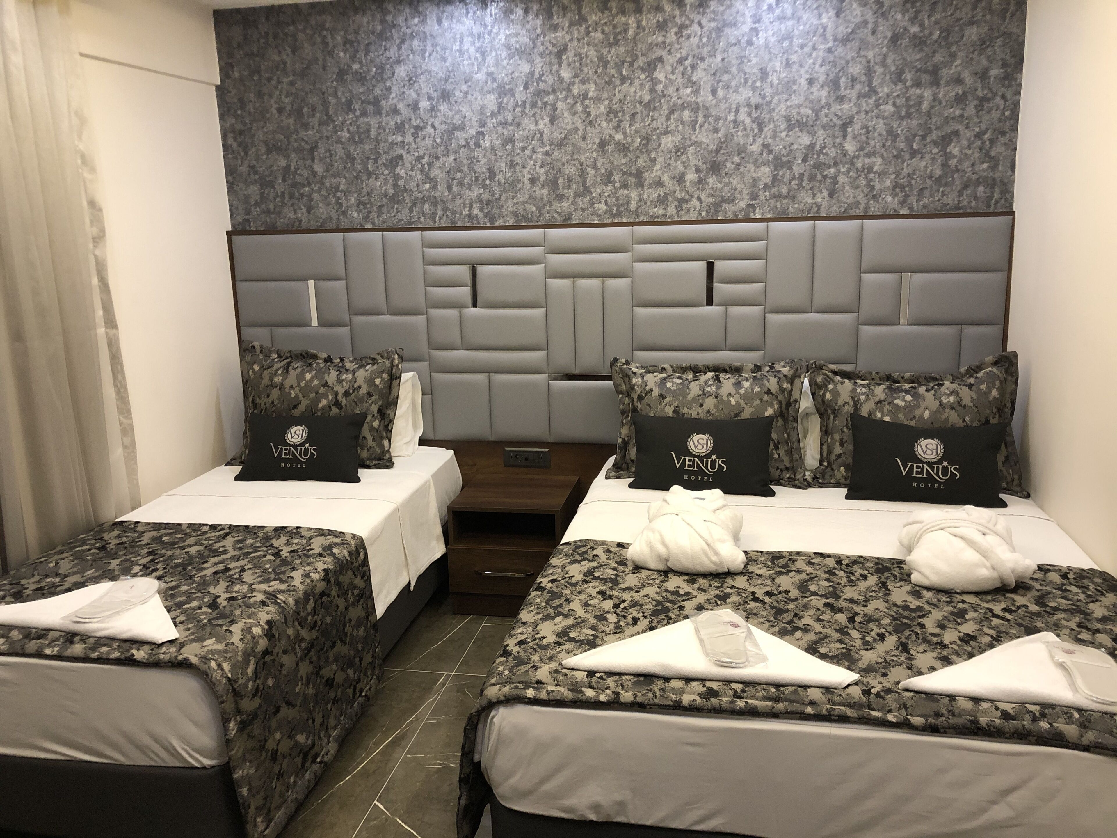 Habitación de hotel con dos camas, cada una con cubrecama y almohadas a cuadros de camuflaje. El cabecero presenta un diseño moderno con acabado gris texturizado.