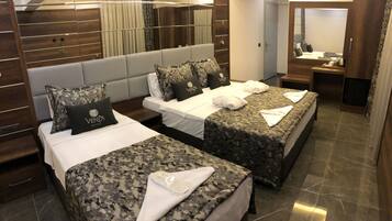 Habitación de hotel con dos camas, cada una con una colcha gris de camuflaje y una almohada con el logo 'Venus Hotel'. La habitación tiene un espejo grande, paneles de madera en las paredes y piso de baldosas.