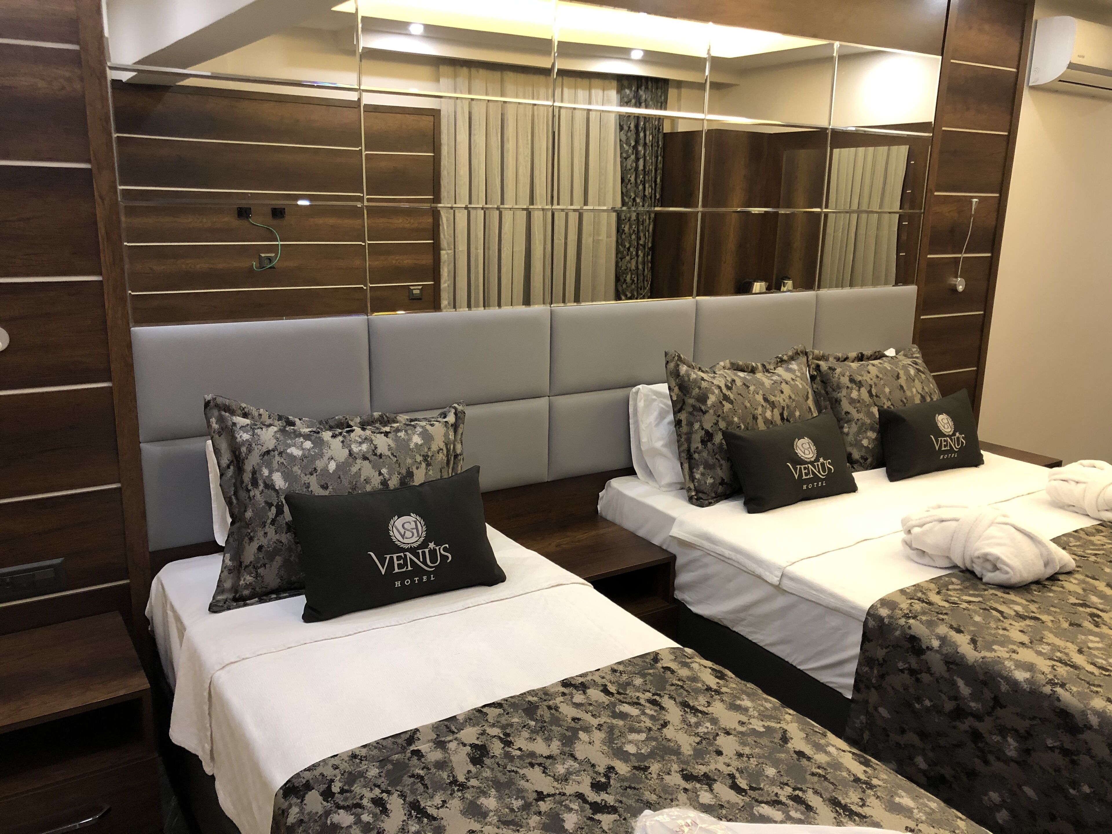 A hotel room with two beds, each with a grey headboard and a camouflage-patterned bedspread. There are matching pillows with the hotel's logo, and a large mirror above the beds.