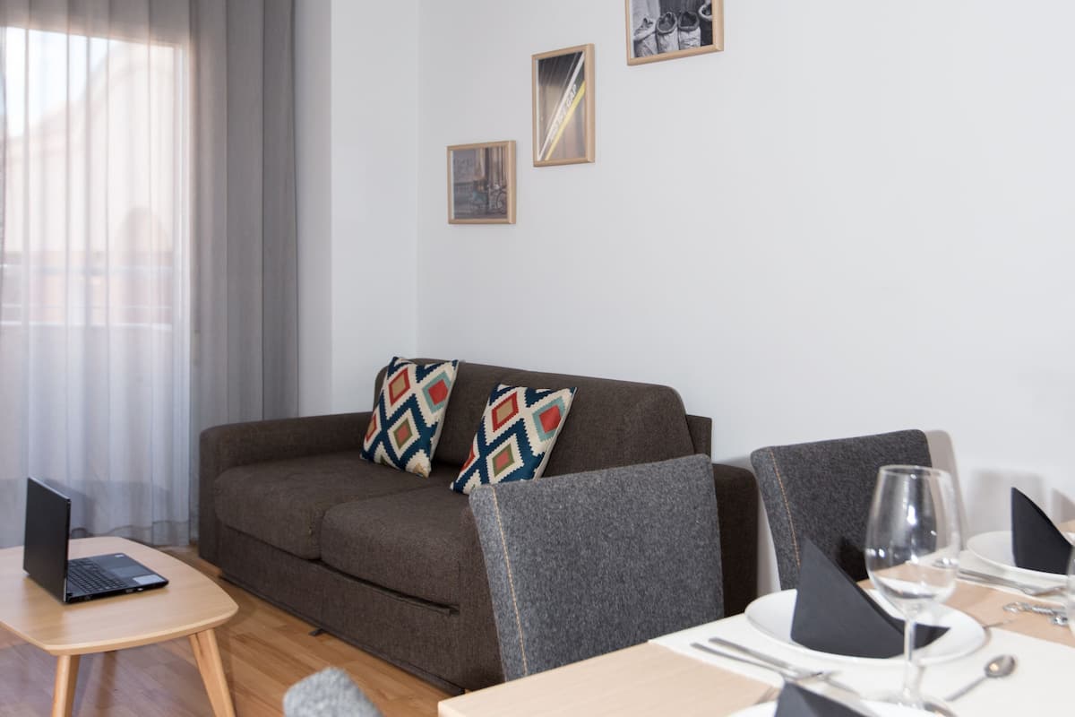 Una sala de estar moderna con un sofá marrón, una mesita de madera con una computadora portátil, y un comedor con sillas y una copa de vino.