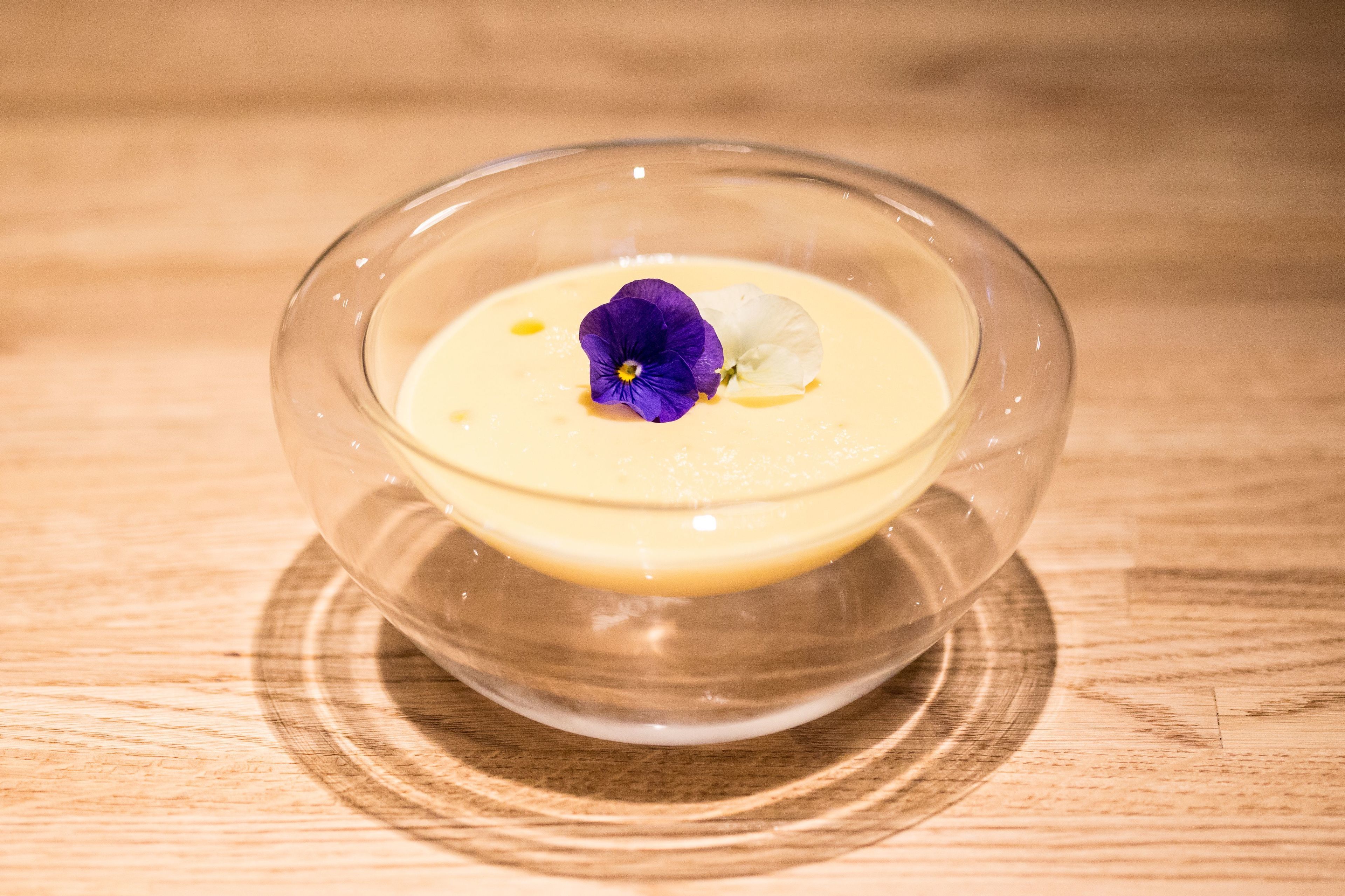 A clear glass bowl with a creamy dessert, a purple flower, and a white flower on a wooden surface.