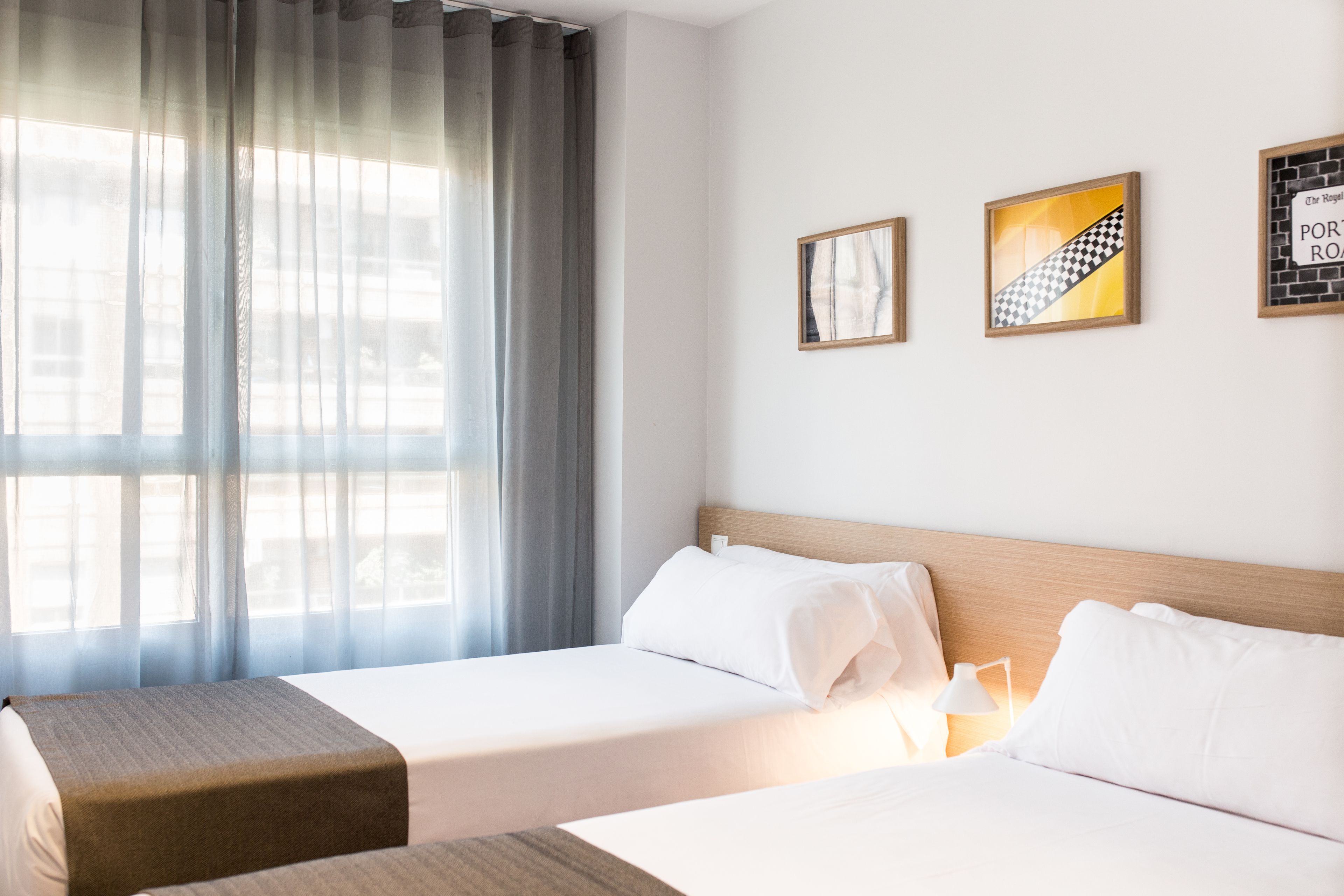 Habitación de hotel con dos camas individuales, un ventanal con cortinas translúcidas y tres cuadros en la pared.