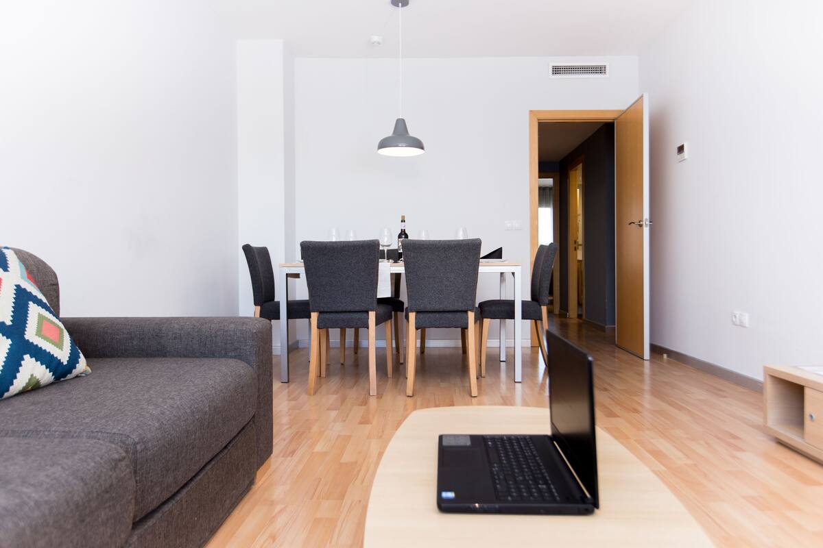 Una sala de estar moderna con un sofá, una laptop sobre una mesa, y un comedor con sillas y una mesa.
