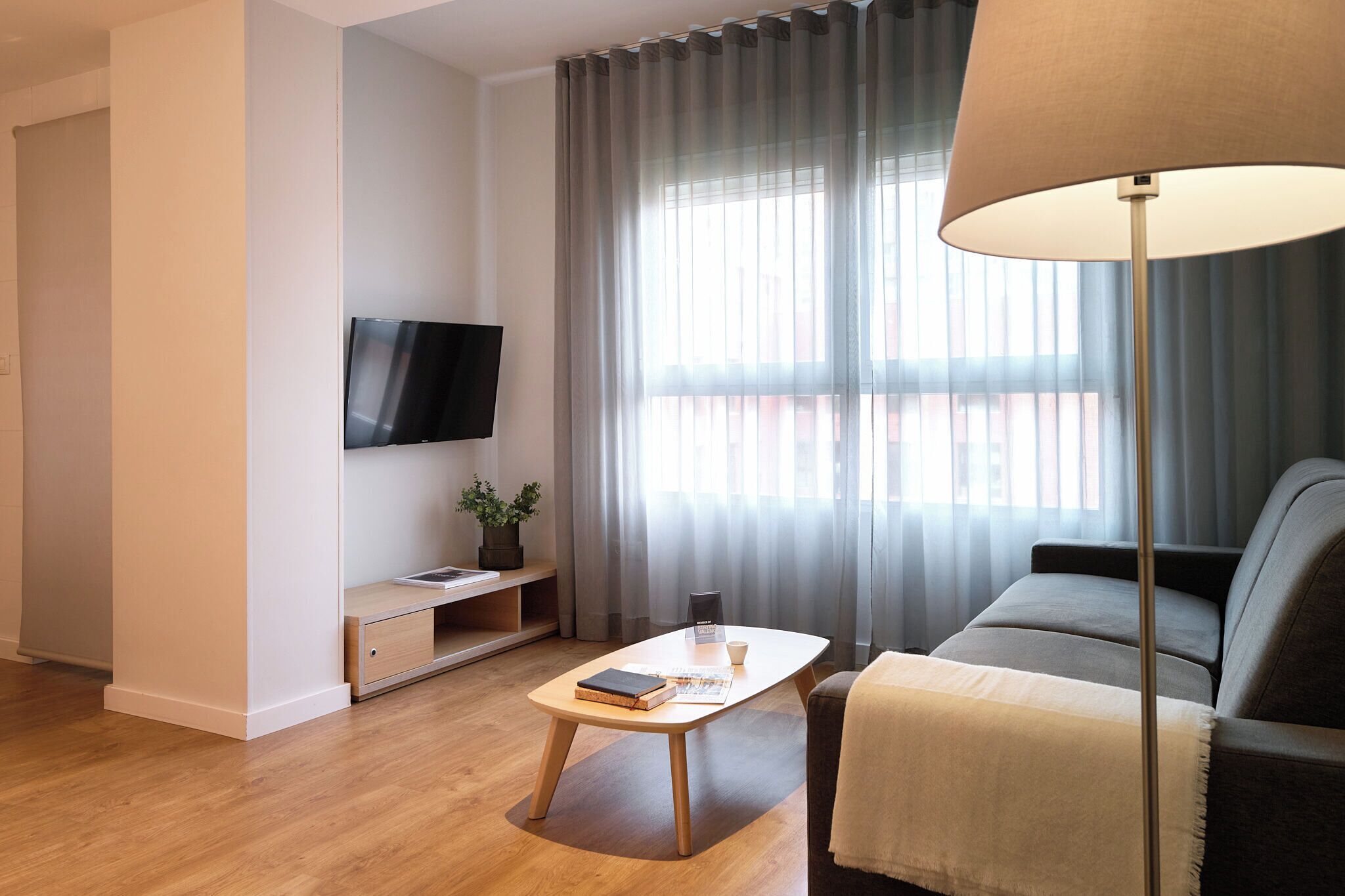 A modern living room with a flat-screen TV mounted on the wall, a wooden coffee table with books and a remote, a sofa with a throw blanket, and a floor lamp.