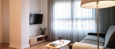 A modern living room with a flat-screen TV mounted on the wall, a wooden coffee table with books and a remote, a sofa with a throw blanket, and a floor lamp.