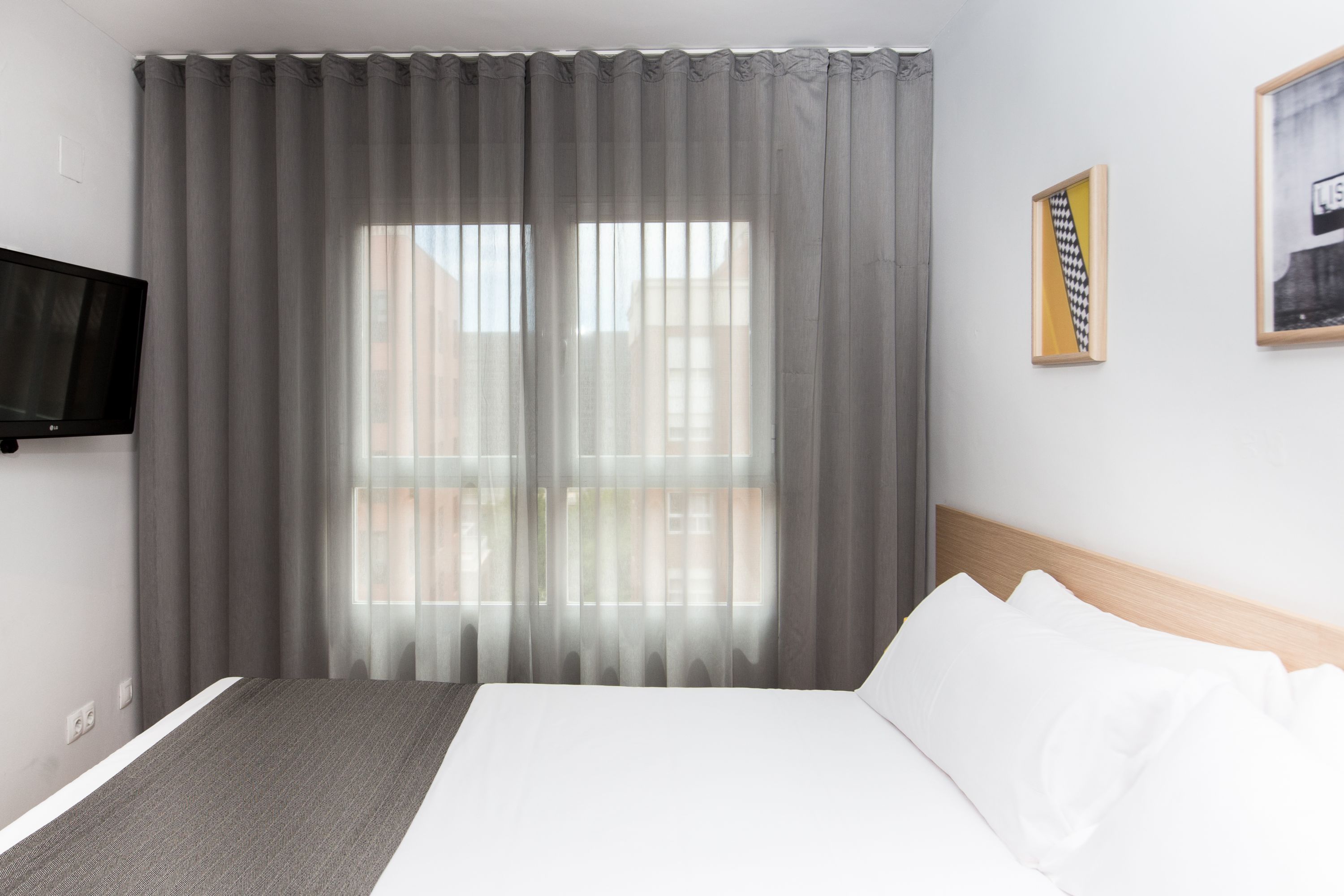 Ein Hotelzimmer mit einem großen Bett, einem flachbildfernseher an der Wand und einem Fenster mit durchsichtigen Vorhängen.