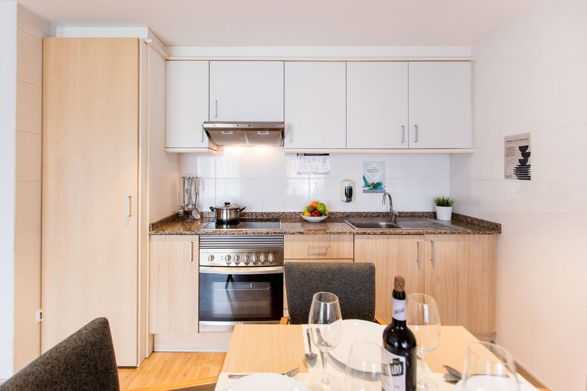 Cocina compacta con armarios blancos, estufa, horno y un comedor con mesa para dos.
