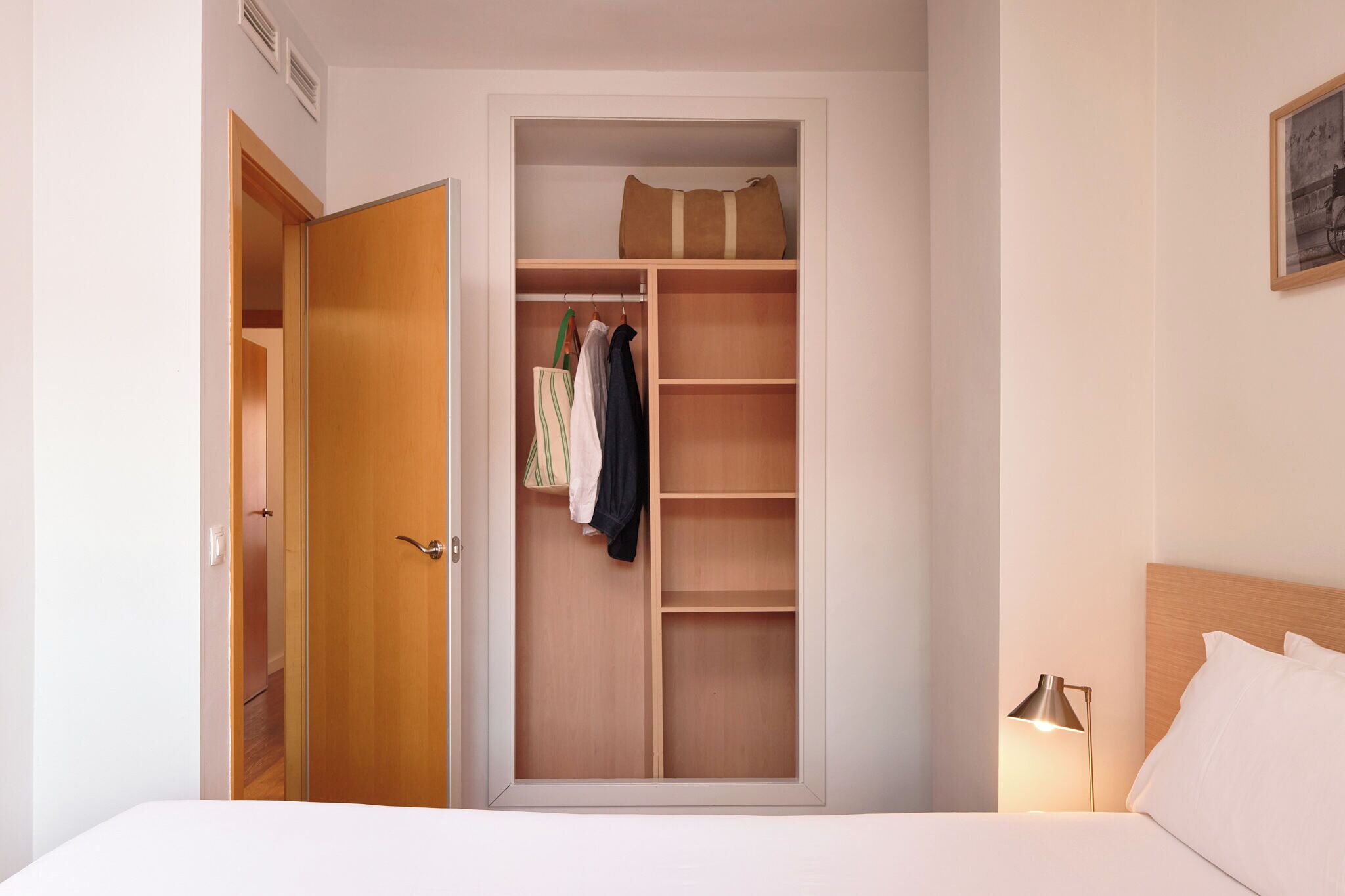 Un dormitorio con un armario de madera, una cama, una lámpara de noche y un cuadro enmarcado en la pared.