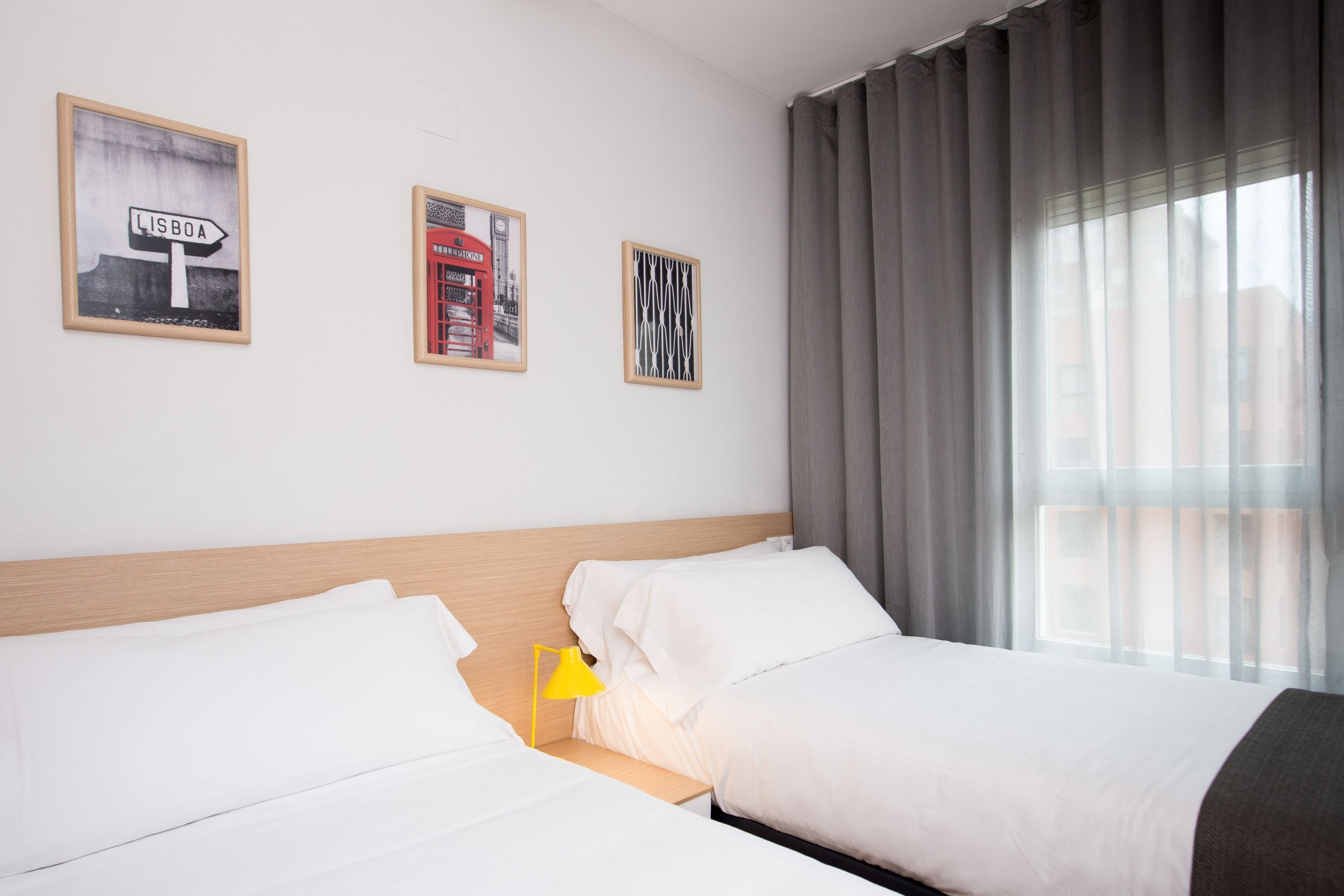 Habitación de hotel con dos camas, cabecero de madera, ventana con cortinas y tres cuadros en la pared.