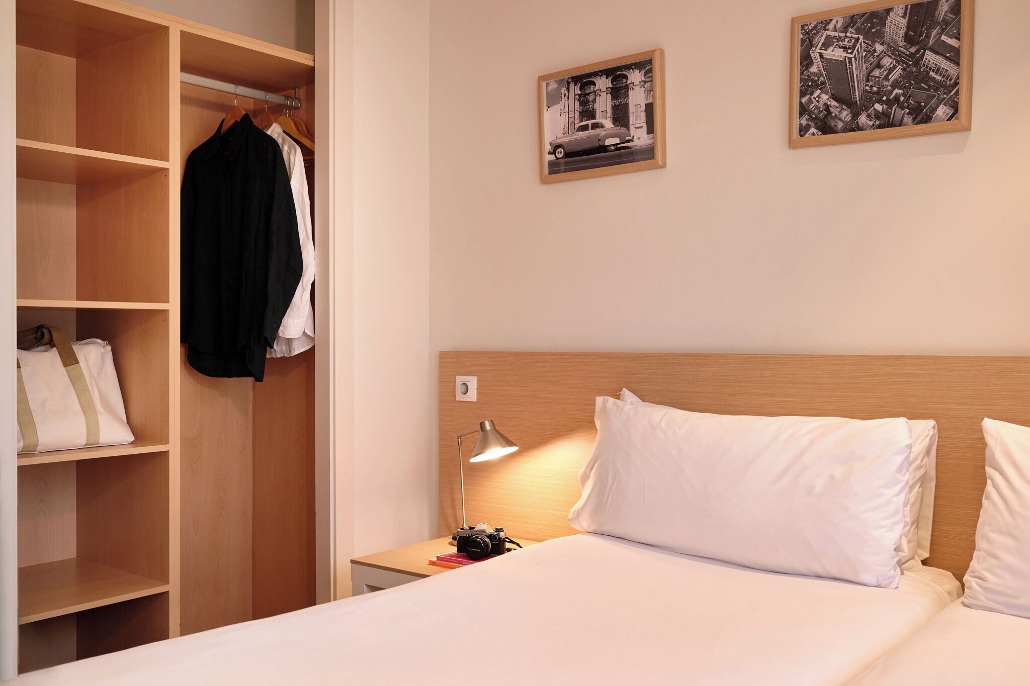 Une chambre d’hôtel bien rangée, avec un lit, une table de chevet, un bureau, une armoire et deux tableaux encadrés accrochés au mur.