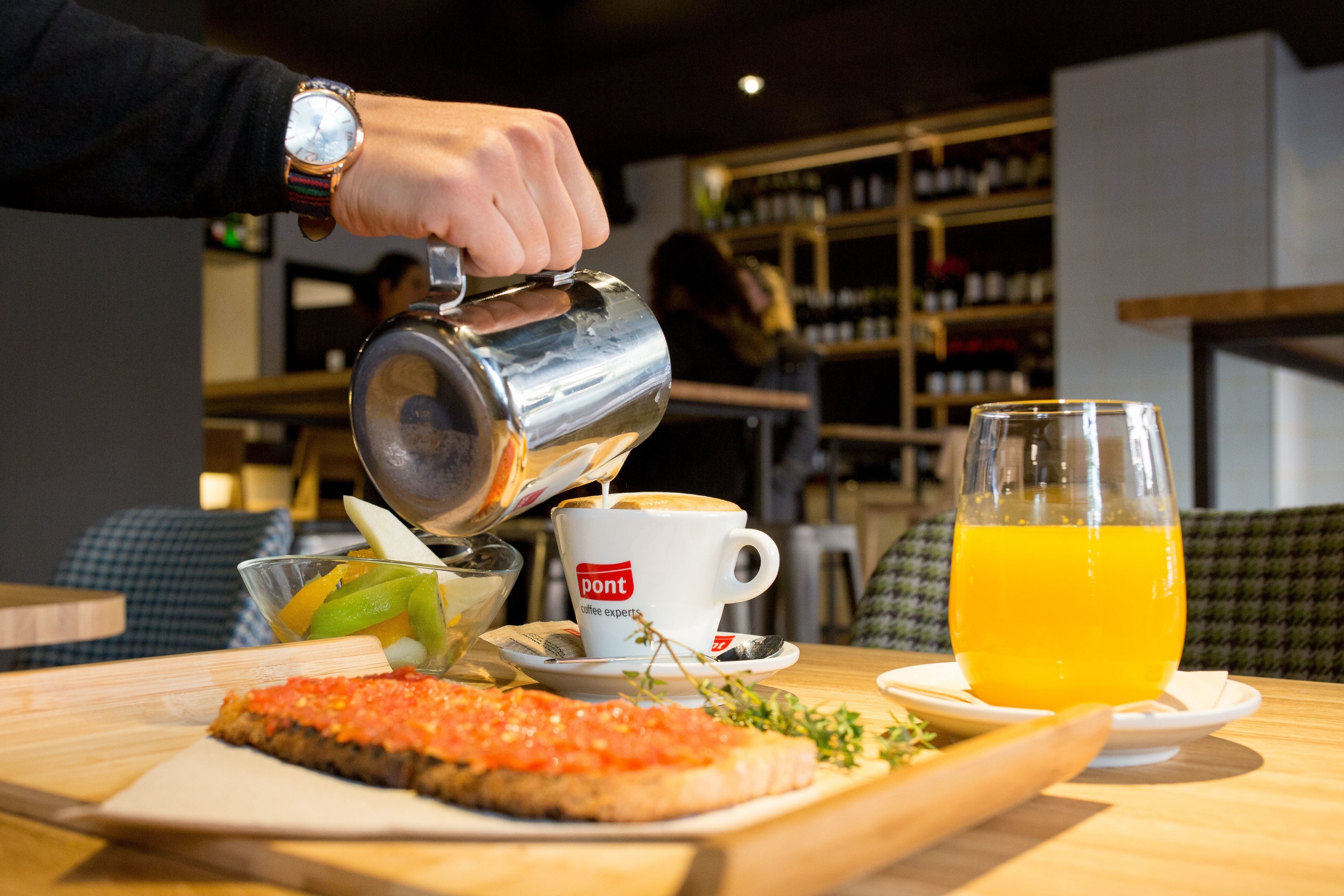 A person pouring a beverage into a cup, a glass of orange juice, a slice of salmon tartare, and a bowl of fruit on a wooden table.