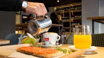A person pouring a beverage into a cup, a glass of orange juice, a slice of salmon tartare, and a bowl of fruit on a wooden table.