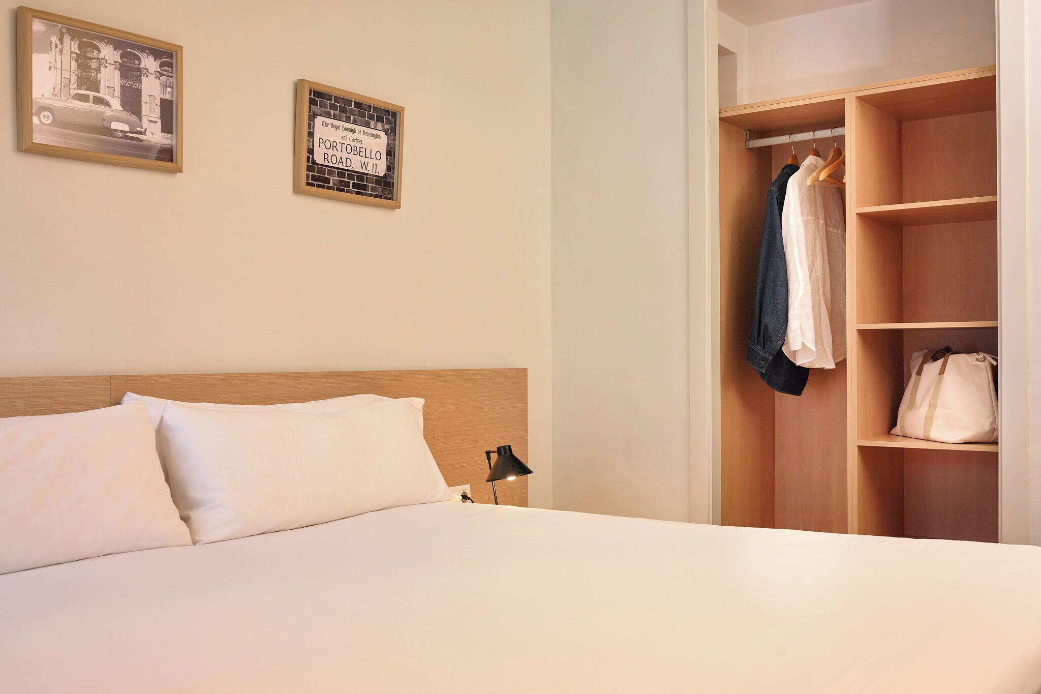 A hotel room with a bed, a nightstand, a lamp, a wardrobe, and two framed pictures on the wall.