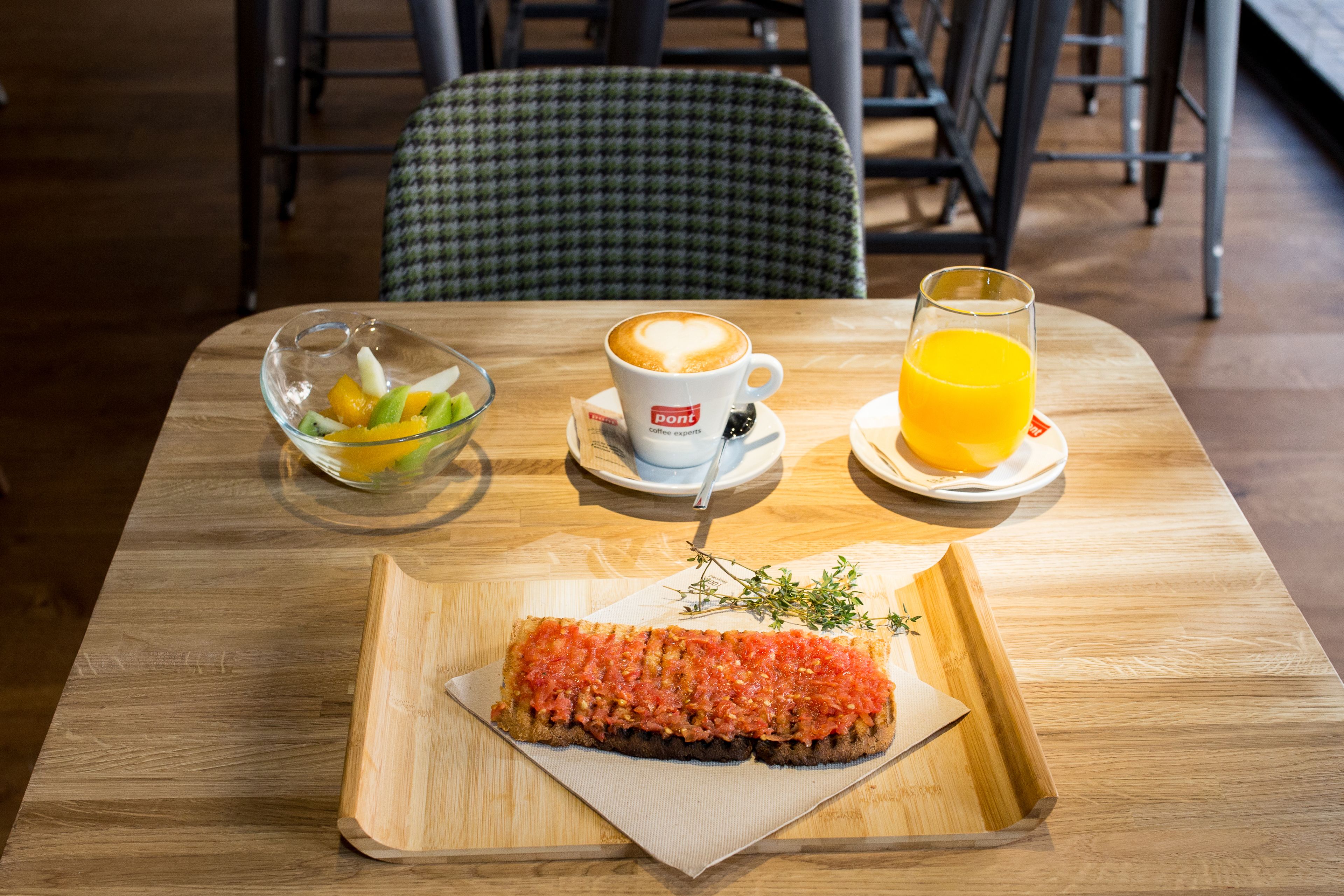 Une table en bois, mise pour le petit-déjeuner, avec un saladier en verre rempli de fruits, une tasse de café, un verre de jus d’orange et une tranche de pain grillé tartinée de purée de tomates.