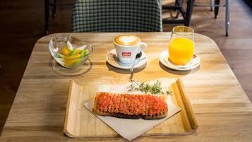 Une table en bois, mise pour le petit-déjeuner, avec un saladier en verre rempli de fruits, une tasse de café, un verre de jus d’orange et une tranche de pain grillé tartinée de purée de tomates.