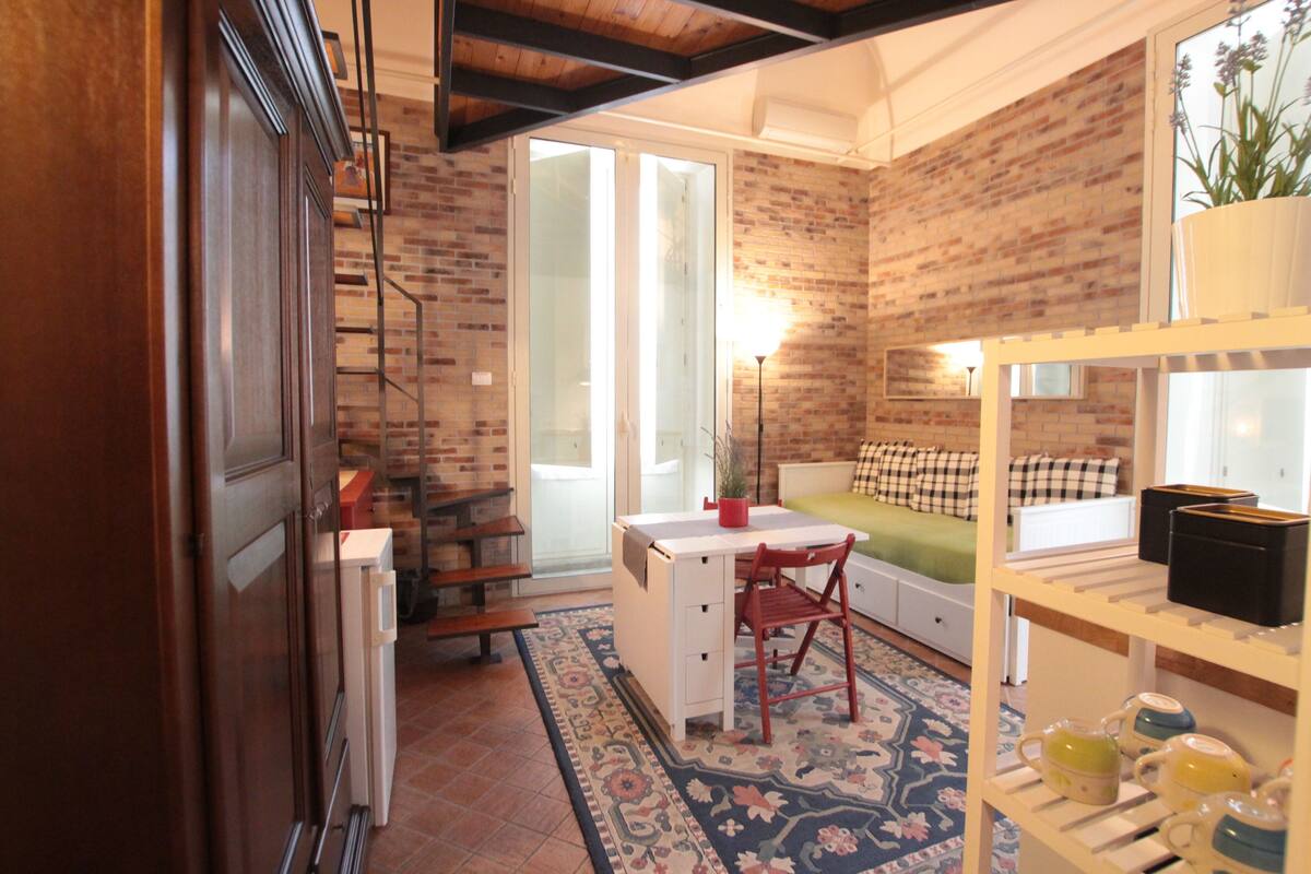 A cozy room with a desk, chair, and shelving unit. There is a staircase leading to an upper level, and a large window providing natural light.