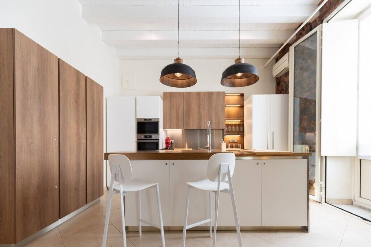 A modern kitchen with wooden cabinets, a central island with bar stools, pendant lighting, and a view through a glass door.